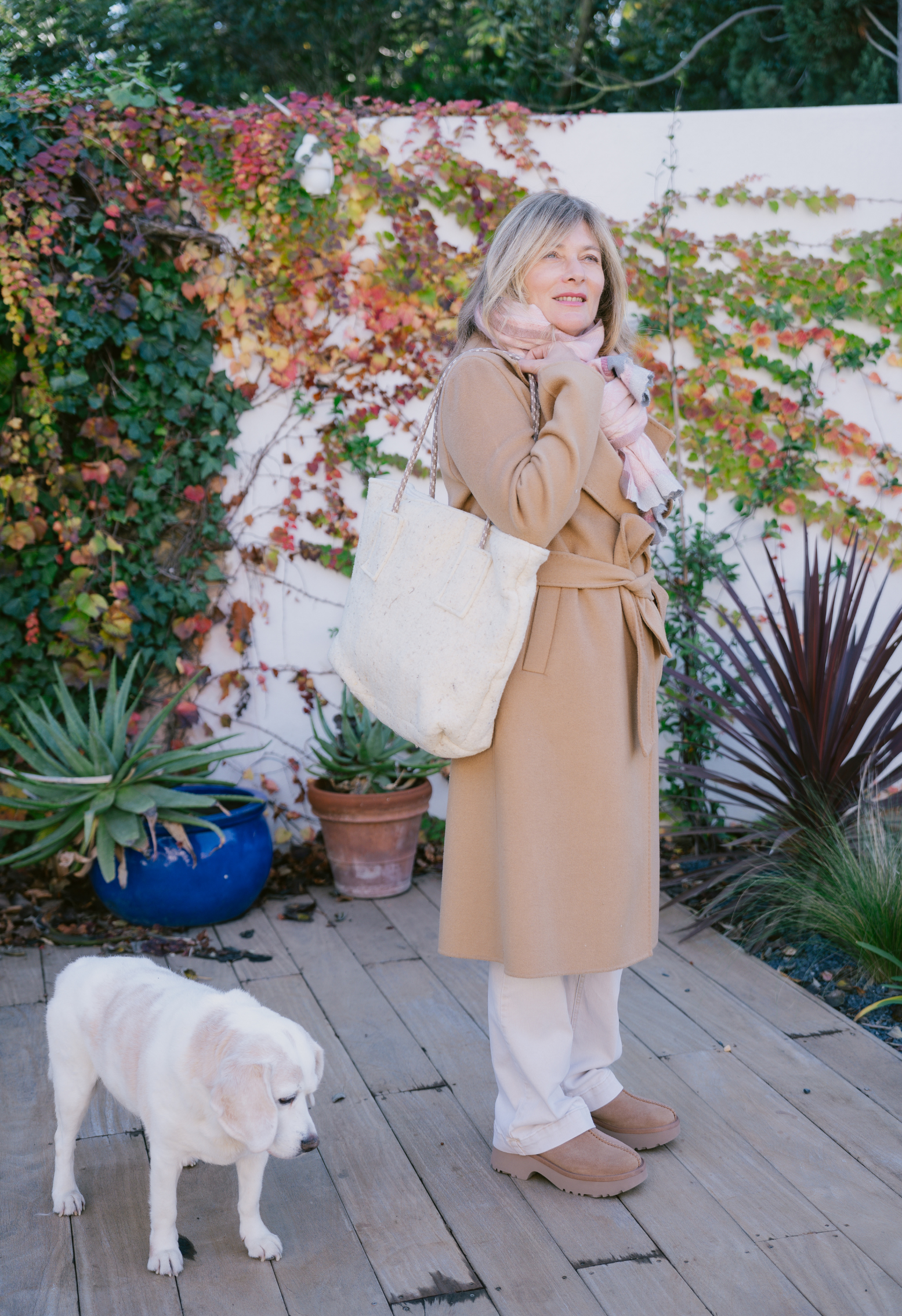 Femme en manteau camel et écharpe rose avec sac en toile, debout devant un mur de lierre, avec un chien blanc.