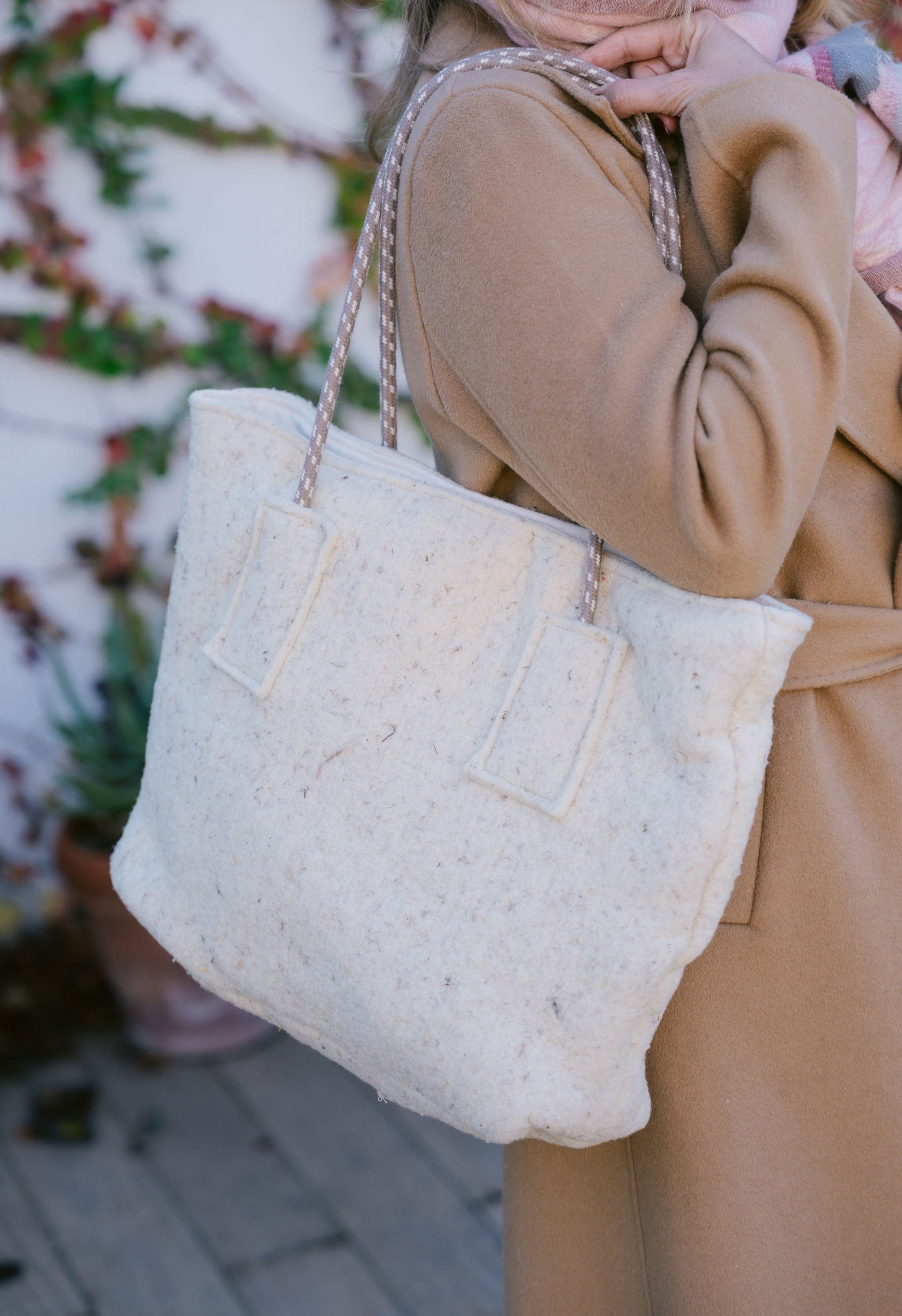 Femme en manteau beige tenant un sac à main en tissu blanc avec des anses en corde torsadée.