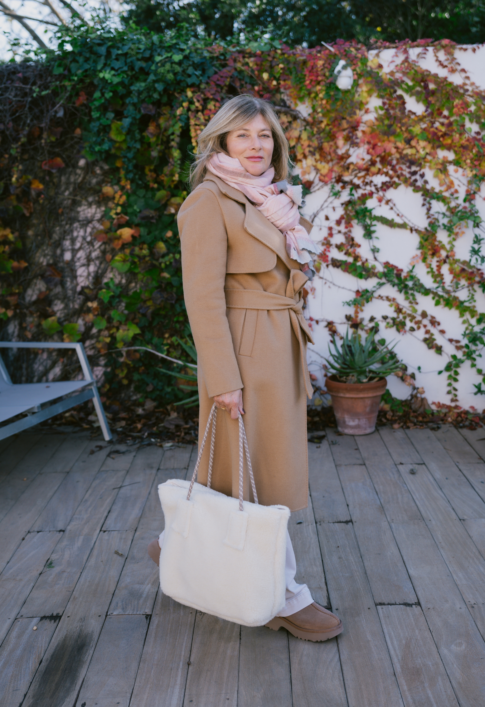 Femme élégante en manteau camel et écharpe rose, tenant un sac blanc. Arrière-plan verdoyant et plantes en pot sur terrasse en bois.