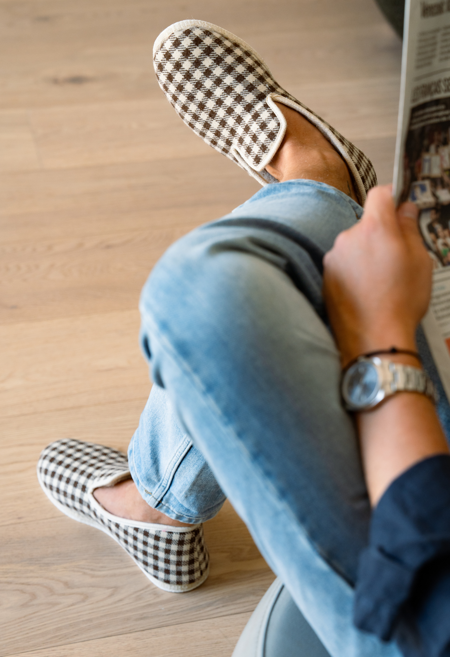 Homme lisant le journal, portant des jeans et des chaussons à carreaux, vue rapprochée sur les pieds, une montre.