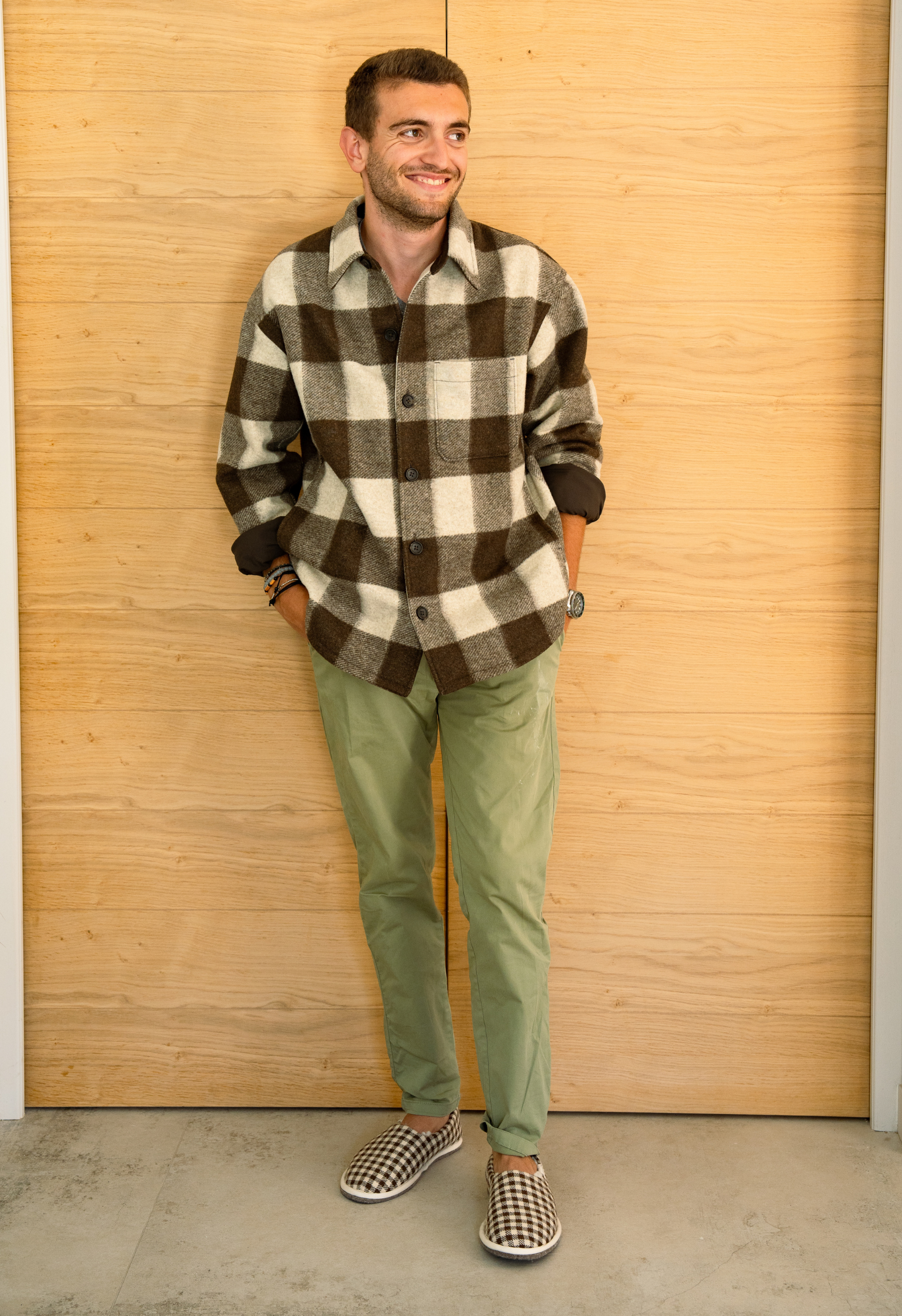 Homme souriant, appuyé contre un mur en bois, vêtu d'une chemise à carreaux marron et beige, un pantalon vert et des pantoufles.