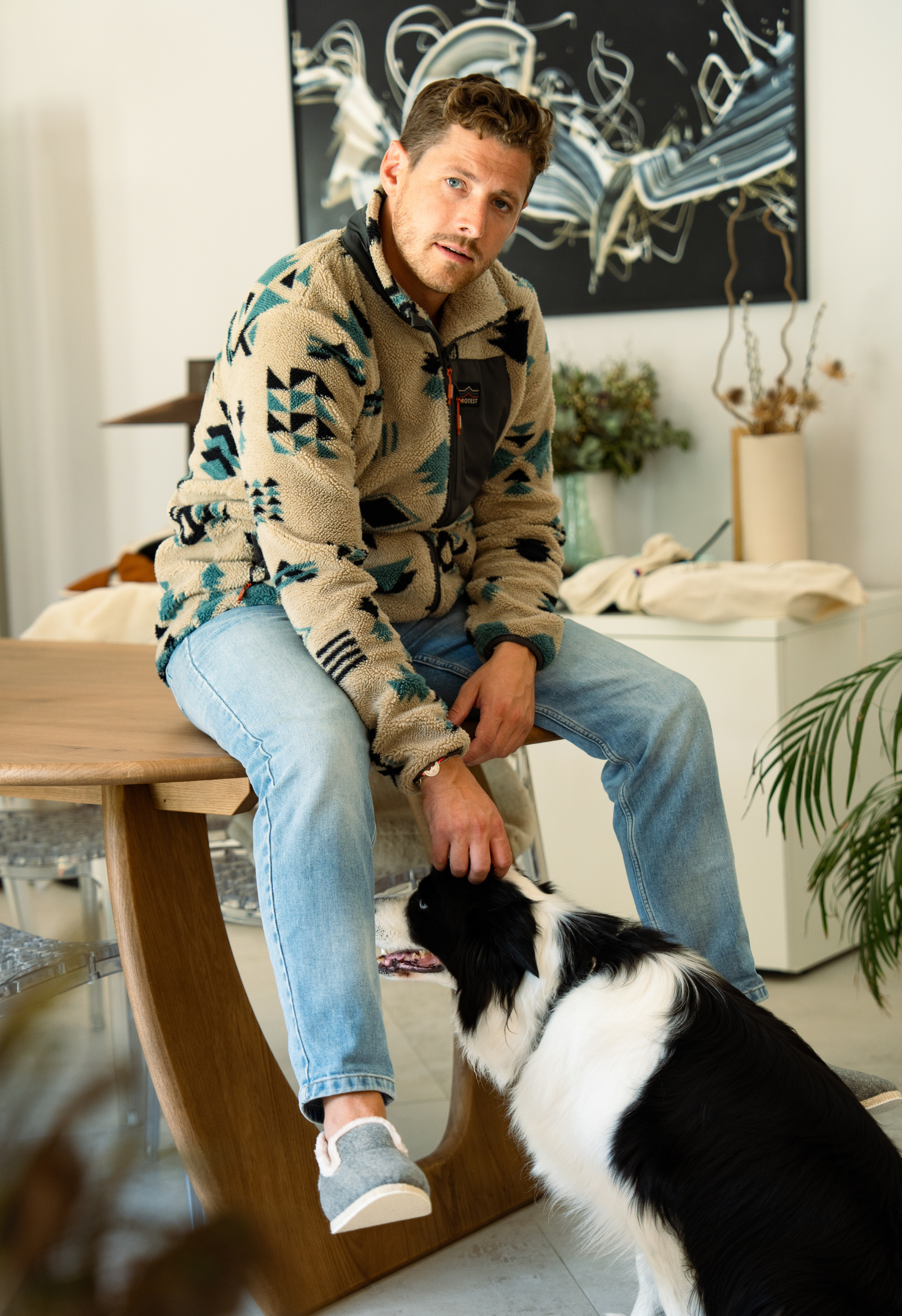 Un homme caresse un chien noir et blanc assis sur une table en bois. L'homme porte une veste à motifs, un jean et des pantoufles.