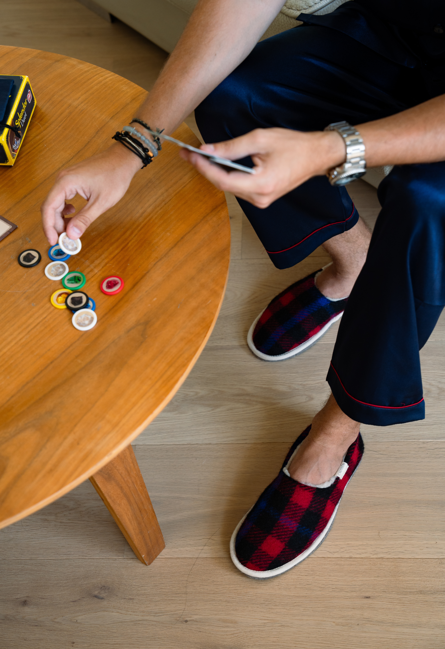 Homme jouant à un jeu de société sur une table en bois. Il porte des pantoufles à carreaux et un pyjama en soie bleu.
