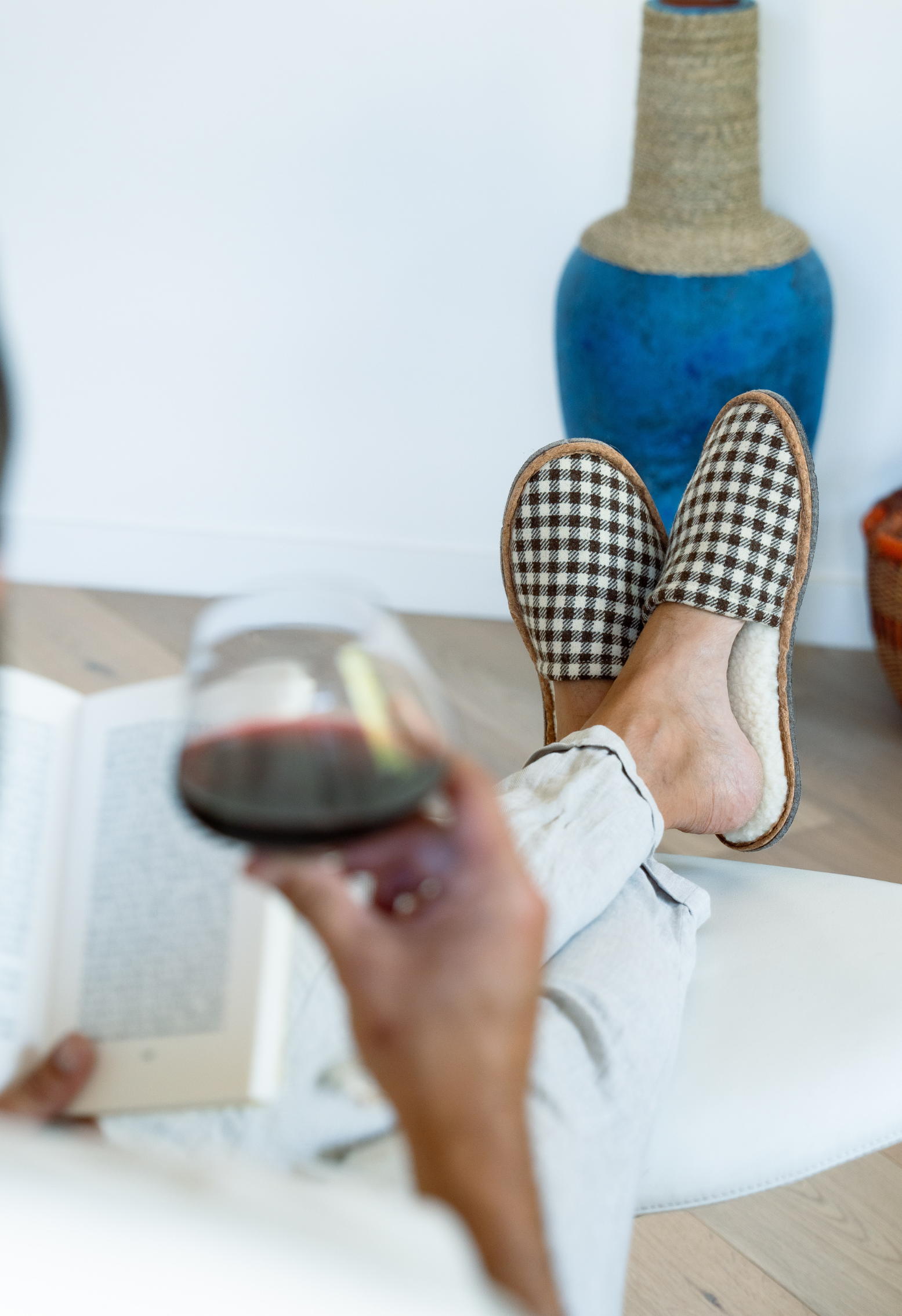 Une personne lit un livre tout en sirotant du vin rouge, les pieds chaussés de pantoufles à carreaux, détente à la maison.