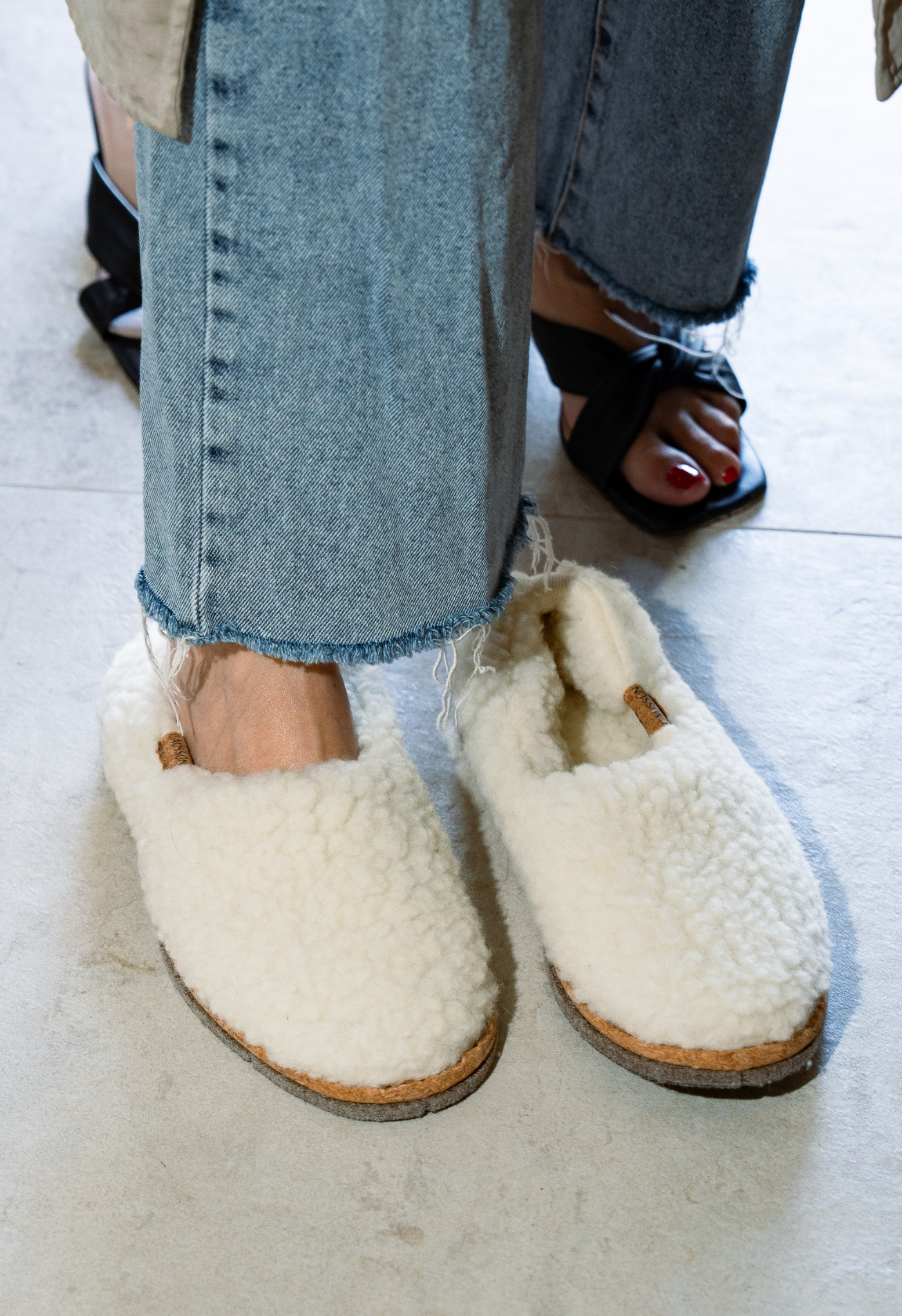 Gros plan de pieds chaussés de pantoufles en laine blanche avec des jeans déchirés, et des sandales noires sur le sol.