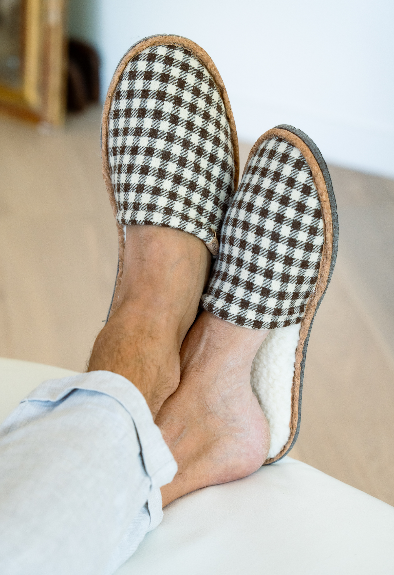 Pieds d'une personne portant des pantoufles à carreaux marron et blanc avec une bordure en liège, détente sur un canapé.