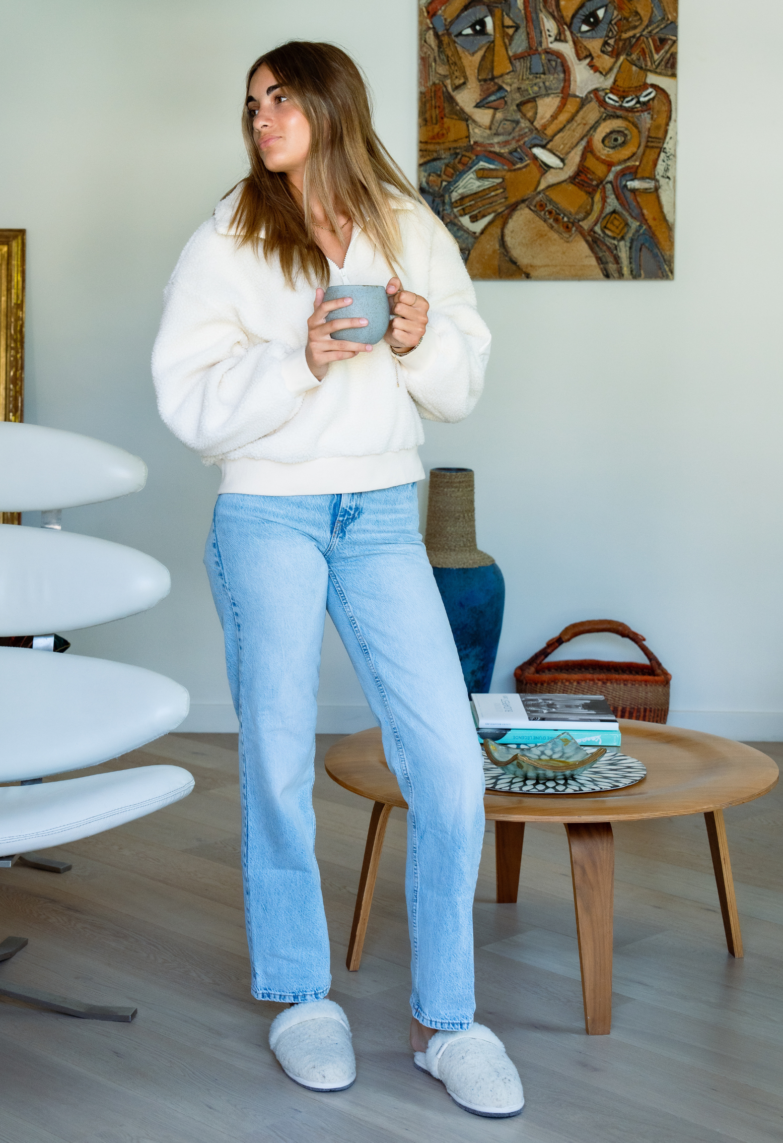 Une jeune femme tenant une tasse de café dans un intérieur moderne. Elle porte un pull en laine, un jean et des chaussons.