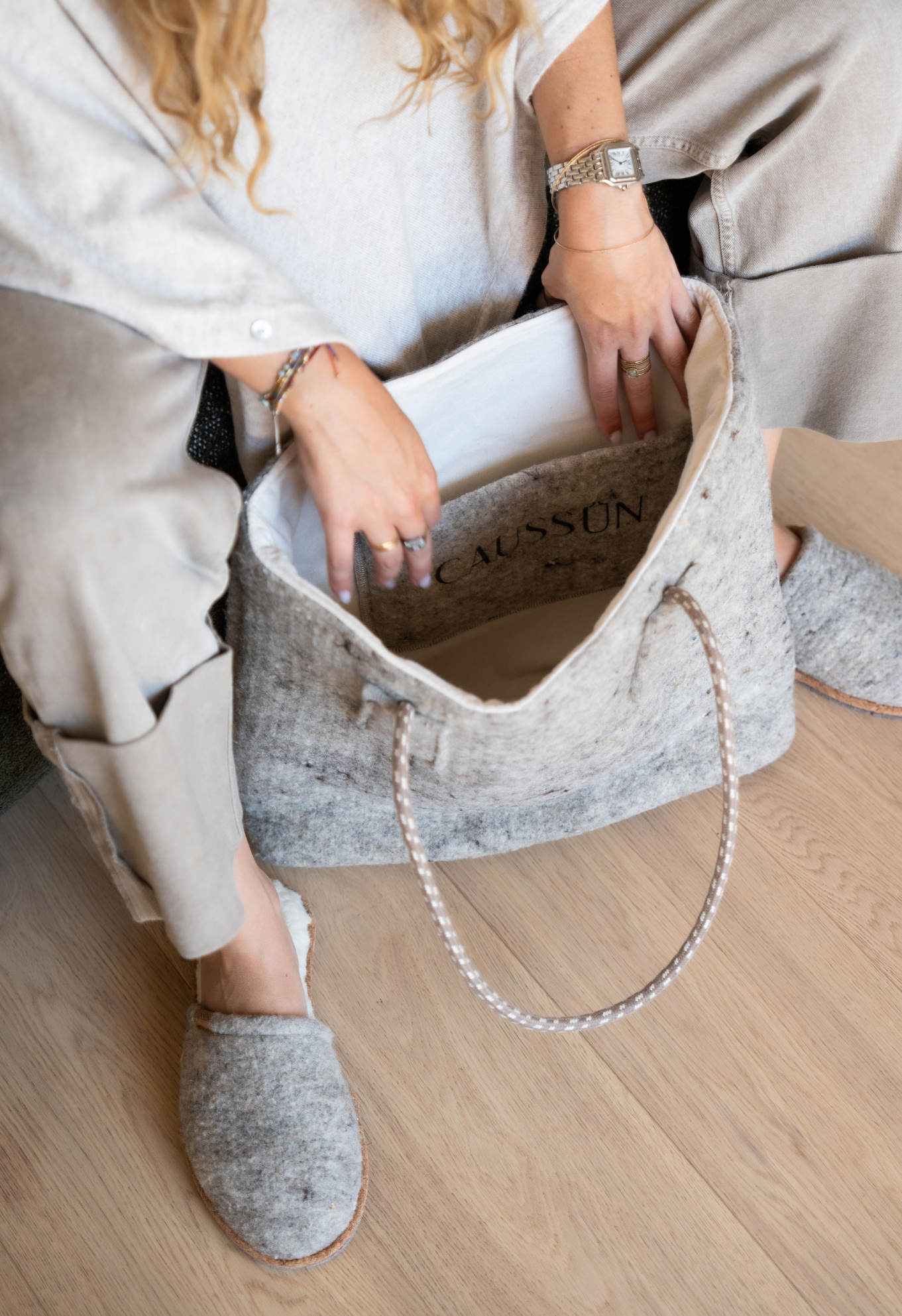 Une femme portant une montre-bracelet, un sac en laine gris avec des chaussons assortis assis sur un sol en bois clair.