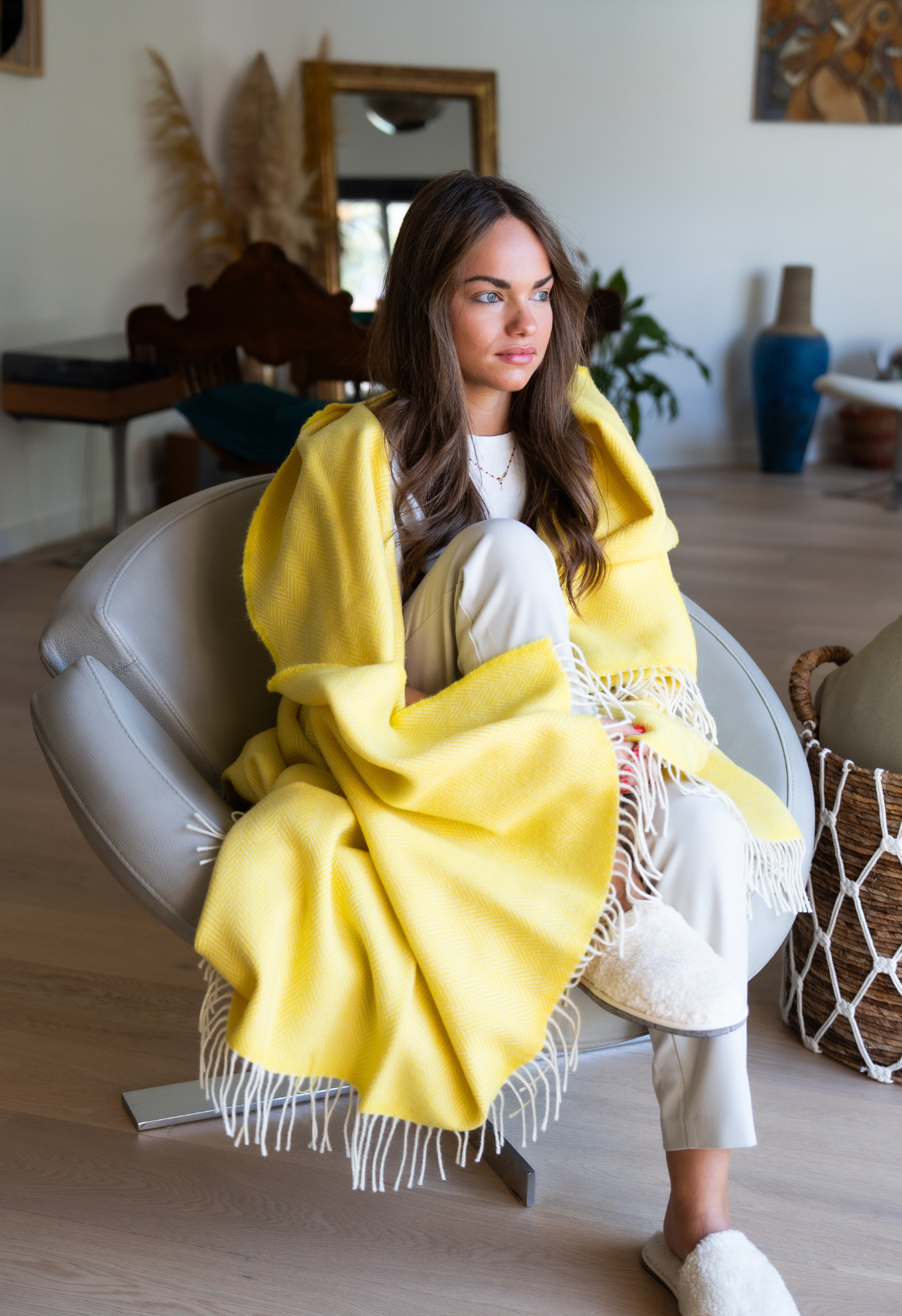 Femme assise dans un fauteuil gris, enveloppée d'une couverture jaune à franges, dans une pièce lumineuse et chaleureuse.