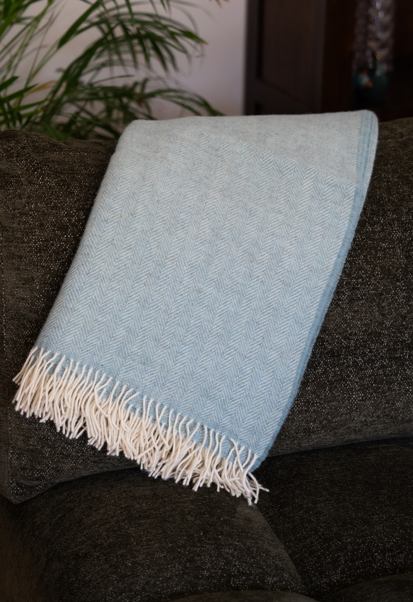 Plaid bleu clair en laine posé sur un canapé sombre. Détails du tissage en chevrons et des franges blanches. Ambiance cocooning.