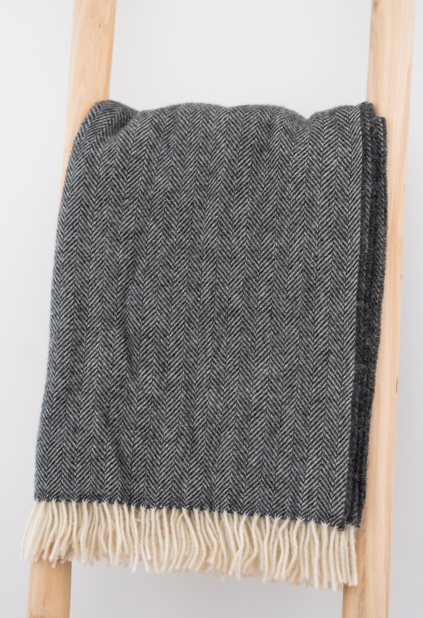 Couverture en laine grise à chevrons avec franges blanches, posée sur une échelle en bois clair.
