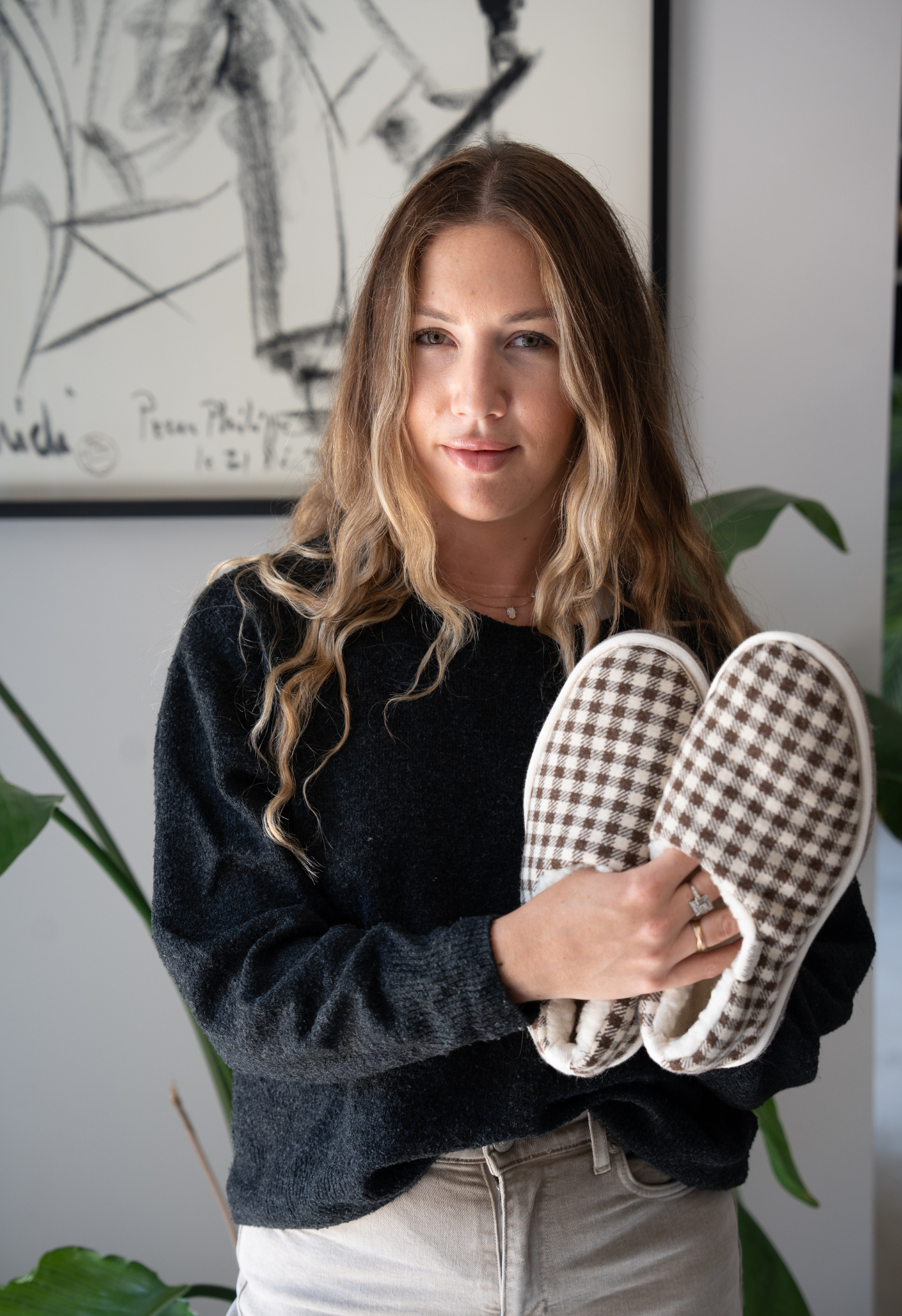 Une femme aux cheveux blonds tient une paire de pantoufles à carreaux dans ses mains. Elle porte un pull noir et un jean beige.