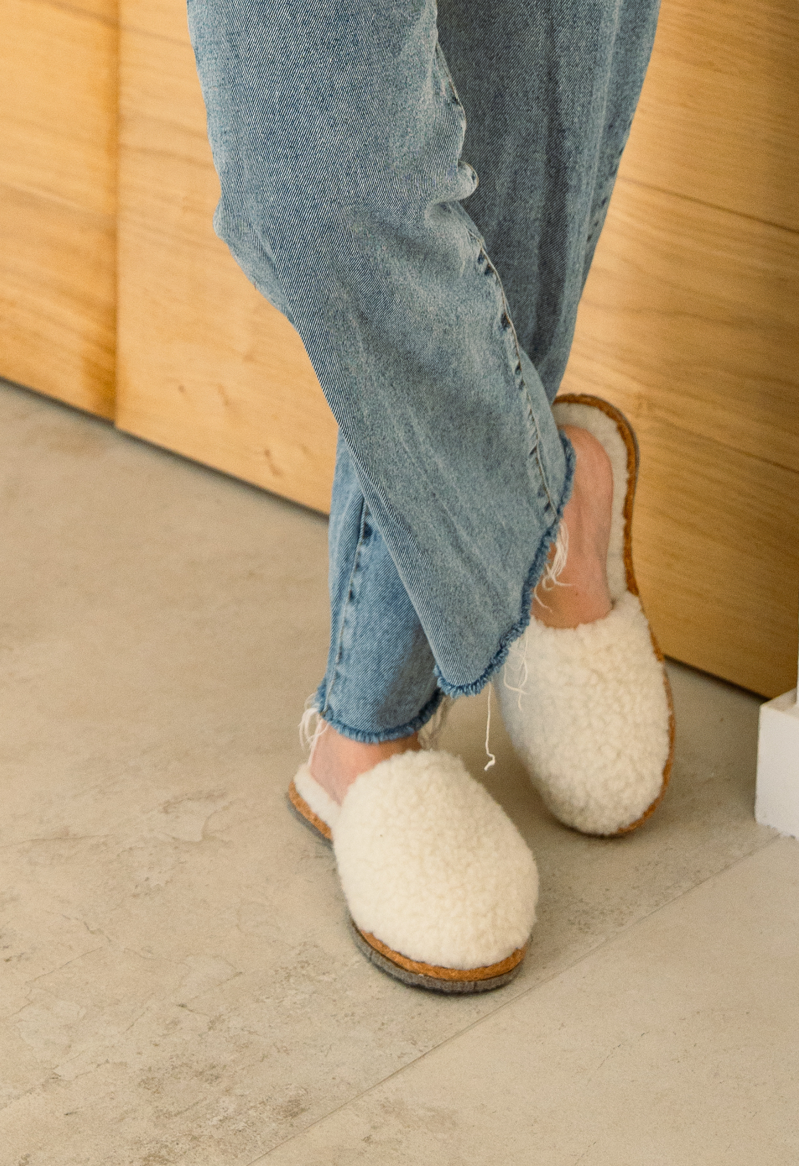 Gros plan des pieds dans des chaussons en laine blanche et un jean bleu clair. Les jambes sont croisées sur le sol clair.
