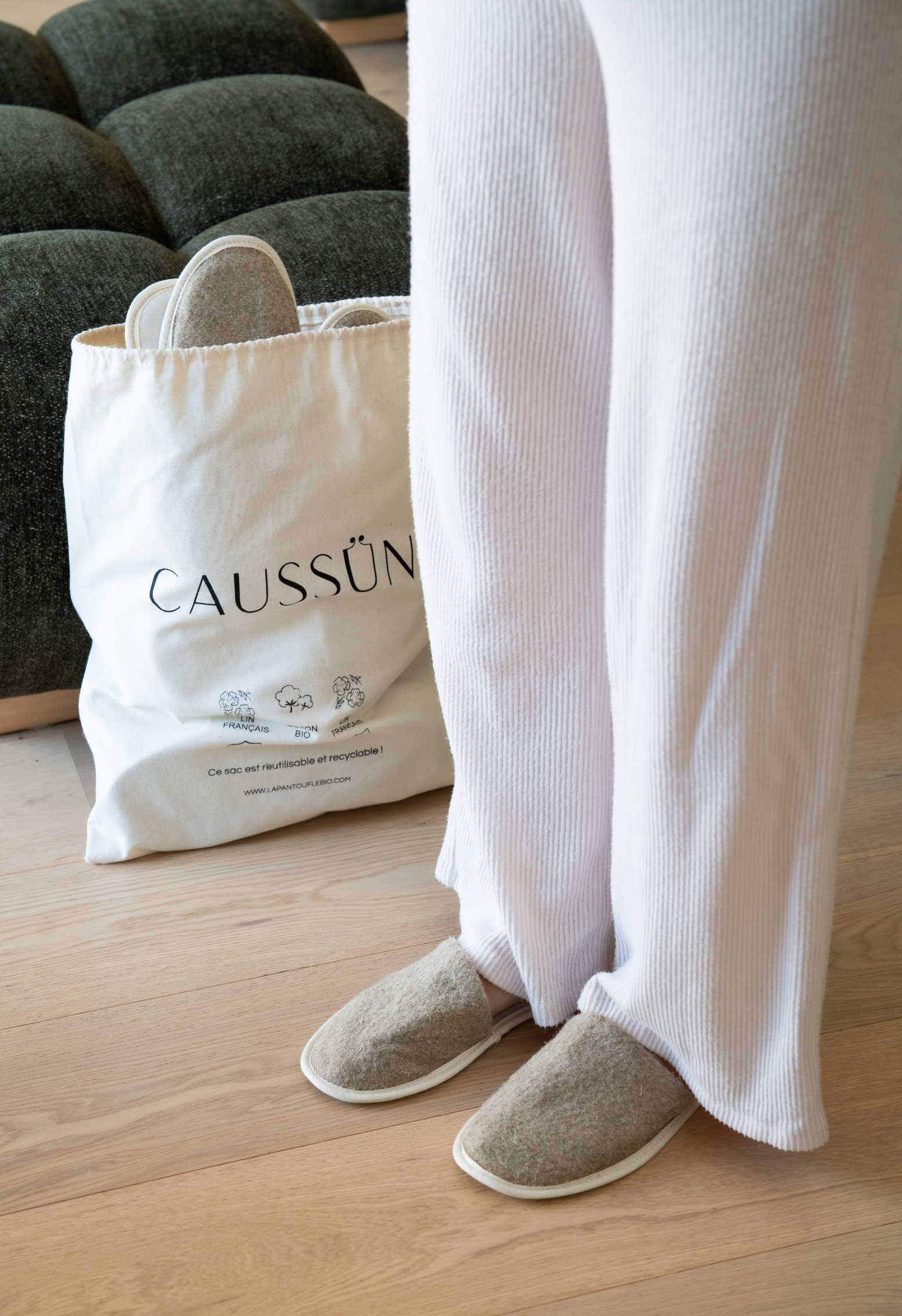 Personne en pantalon blanc et chaussons en laine devant un sac de courses Caussün avec pantoufles.