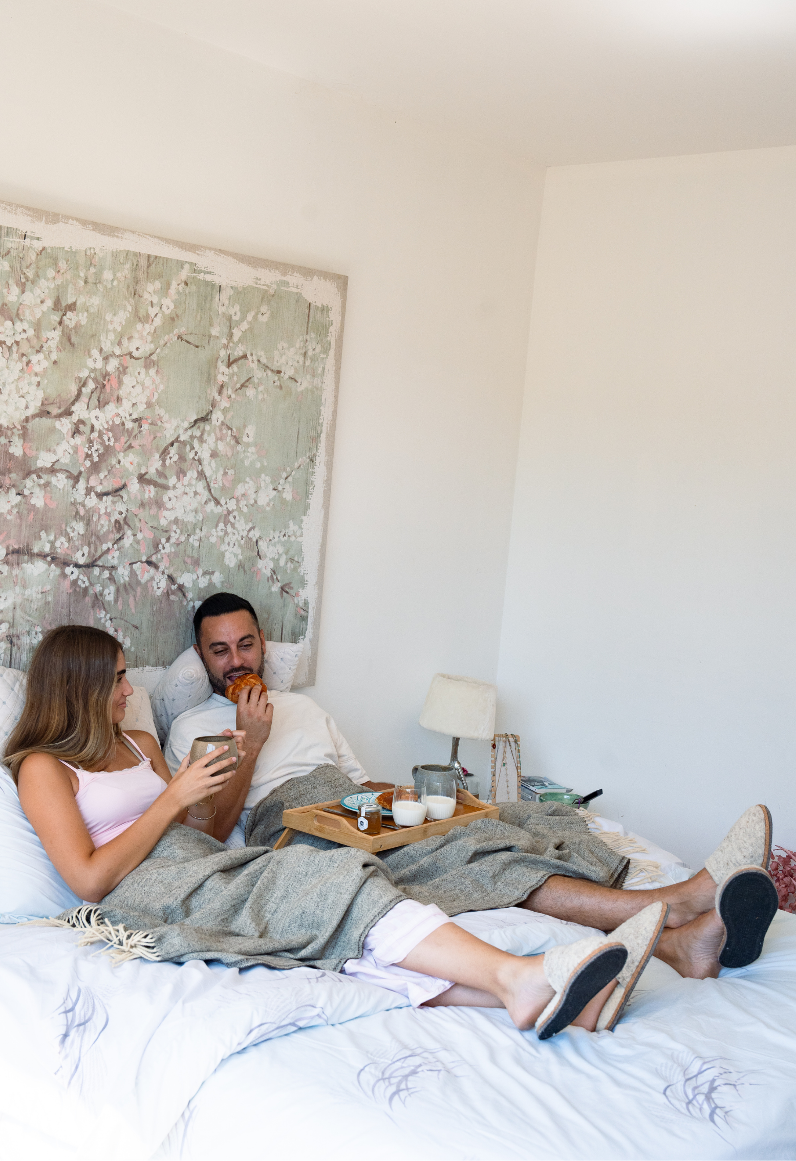 Couple prenant le petit-déjeuner au lit avec un croissant, du café et des chaussons. La chambre est décorée avec une peinture de fleurs.