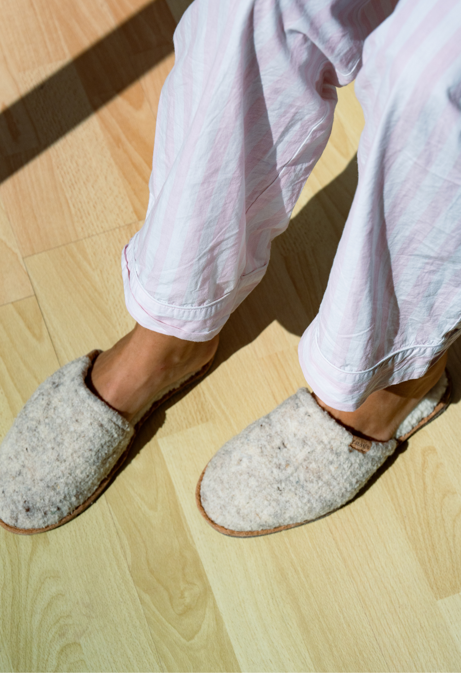Pieds chaussés de pantoufles en laine et d'un pyjama rayé, posés sur un parquet en bois clair, éclairage solaire.