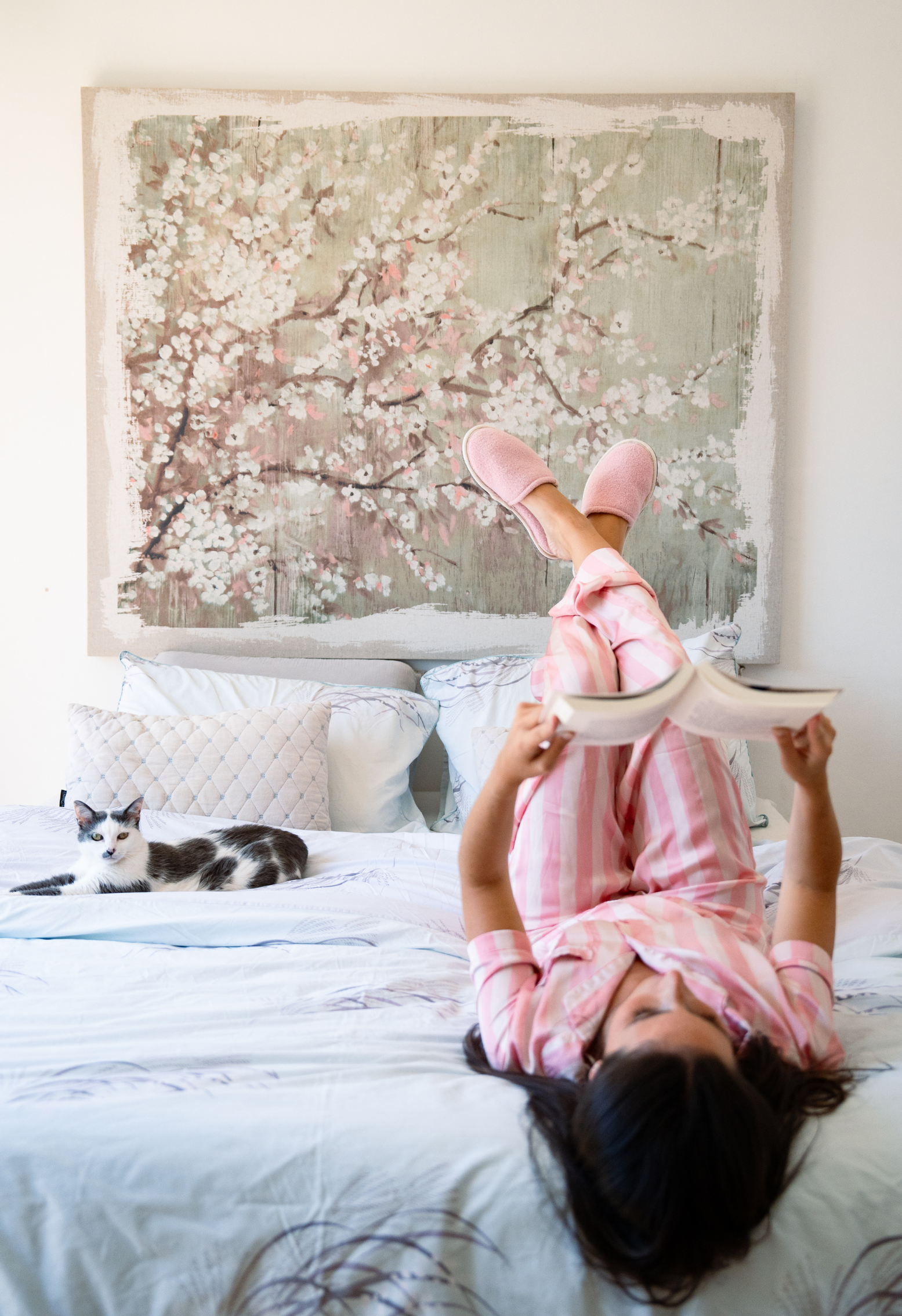 Femme lisant au lit à l'envers, pantalon rose à rayures, chaussons roses, chat et peinture murale de fleurs.