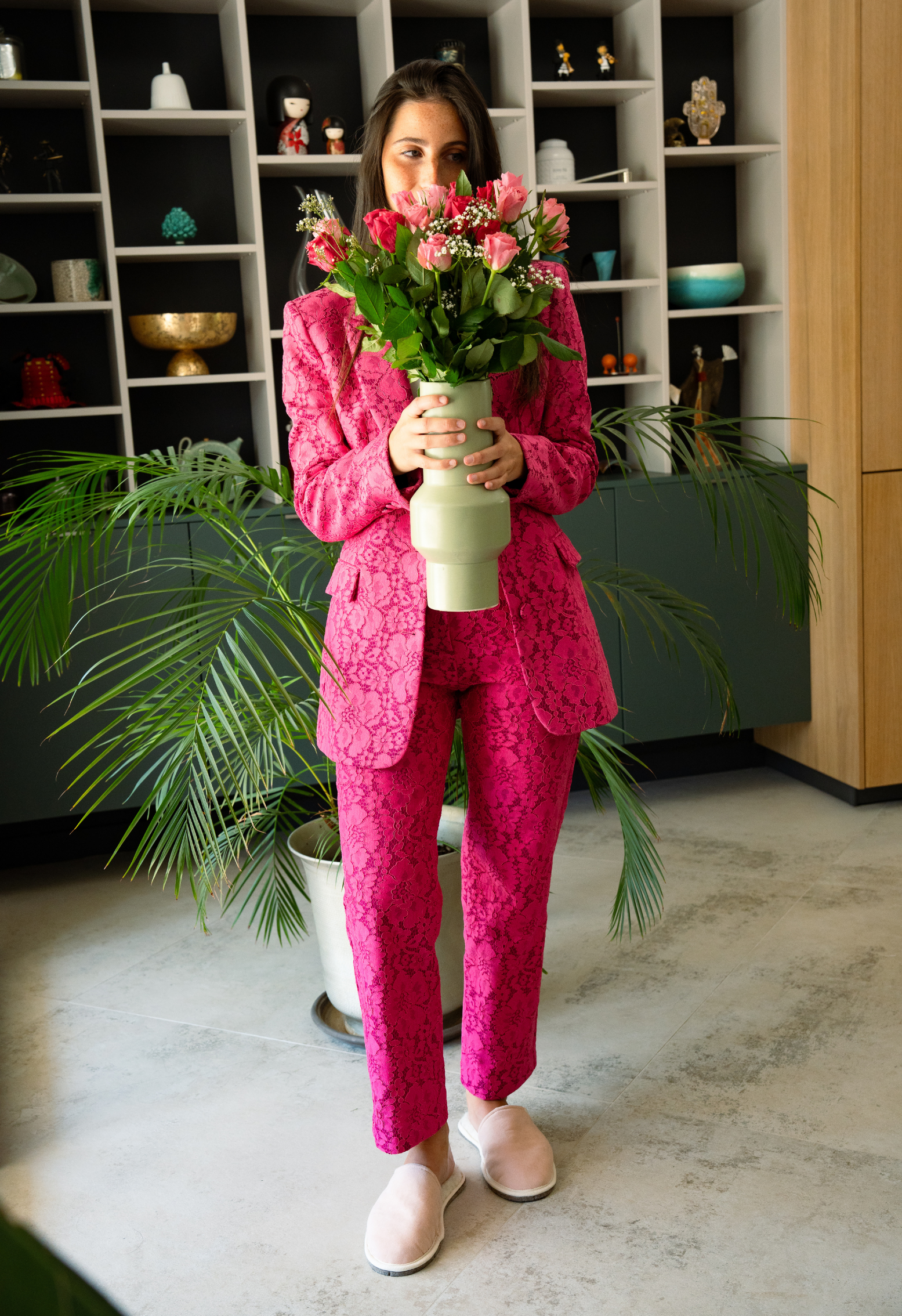 Femme en tailleur rose avec un bouquet de roses devant un meuble avec étagères.