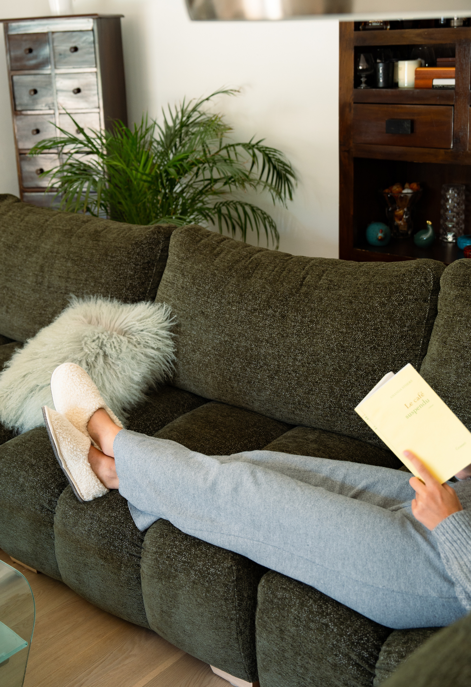 Personne se relaxant sur un canapé vert avec un livre. Les pieds sont recouverts de pantoufles, un coussin moelleux est à côté.