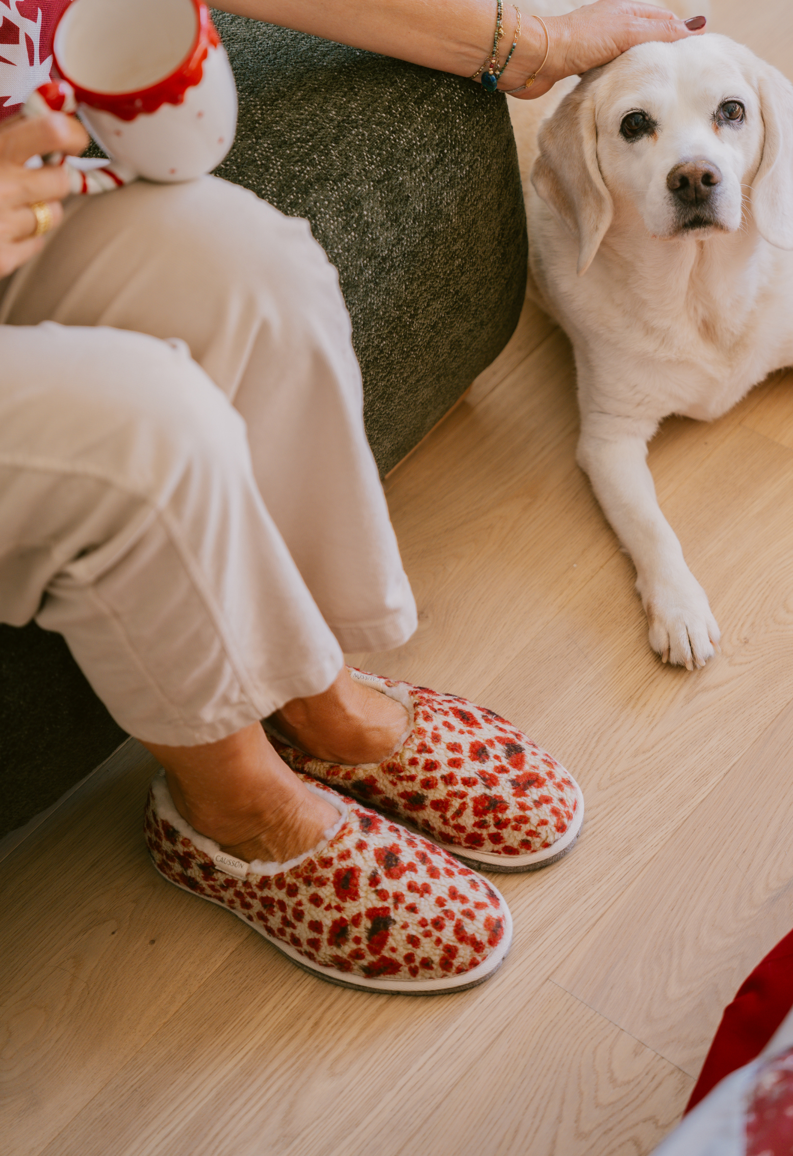 Une personne assise dans son canapé avec un mug et des pantoufles léopard caresse un chien blanc.
