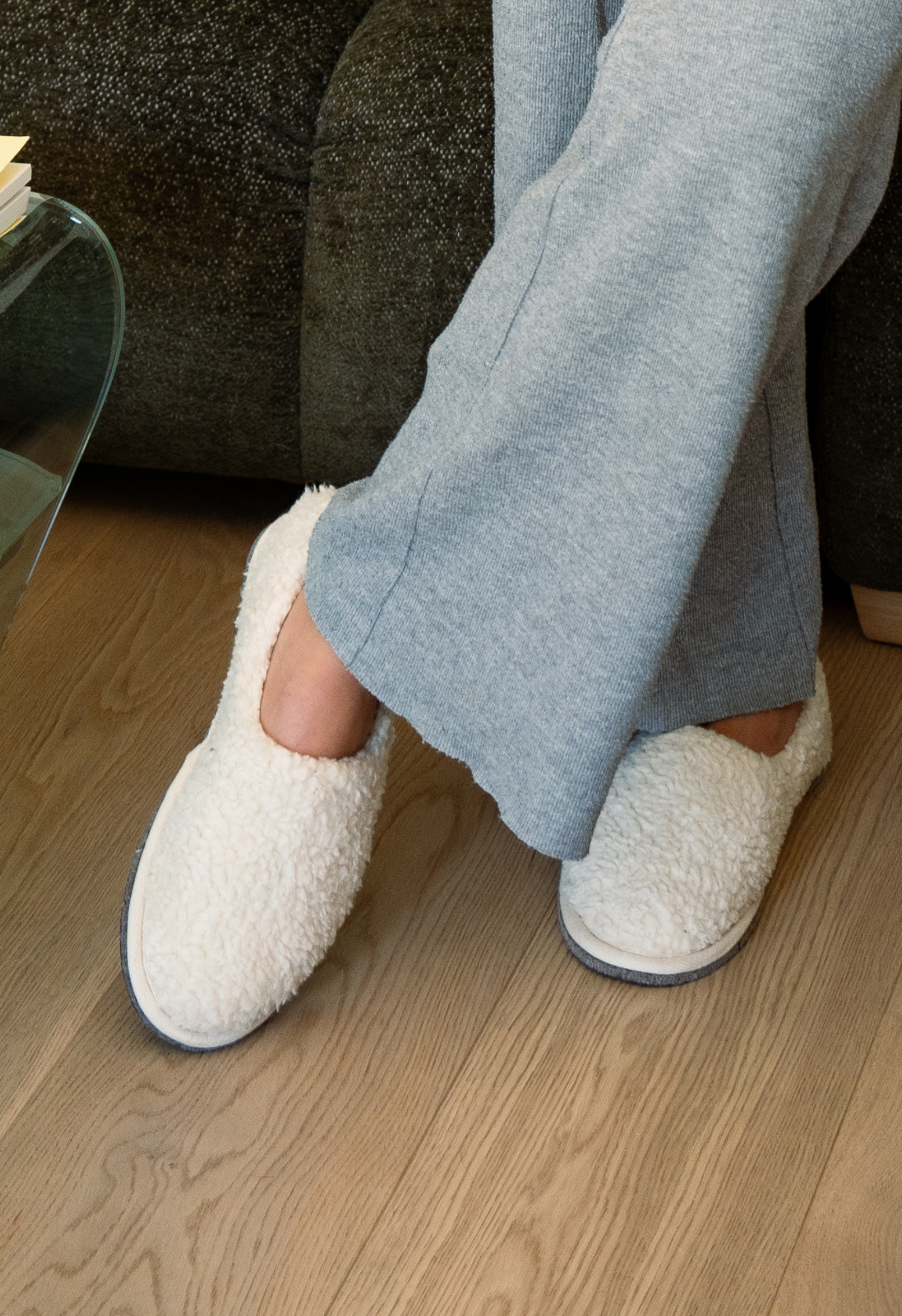 Une personne en pantalon gris et pantoufles blanches en fourrure est assise sur un canapé, les jambes croisées.