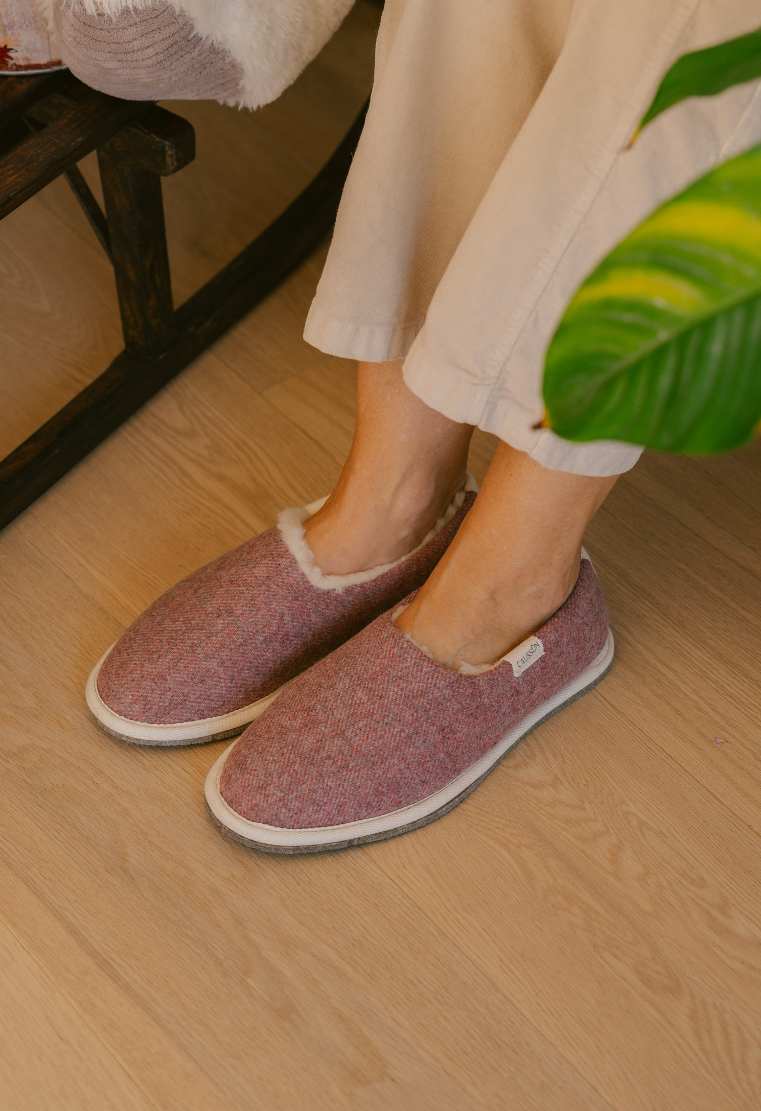 Une personne portant des chaussons en laine rose sur un sol en bois clair. Les chaussons sont doublés de fourrure blanche.