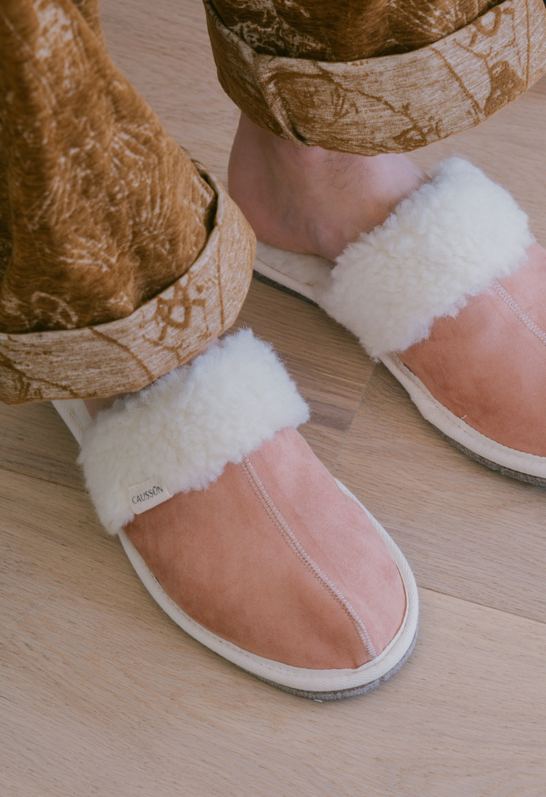 Gros plan des pantoufles roses chaudes avec de la fourrure blanche et des jambes dans un pantalon marron à motifs sur plancher de bois clair.