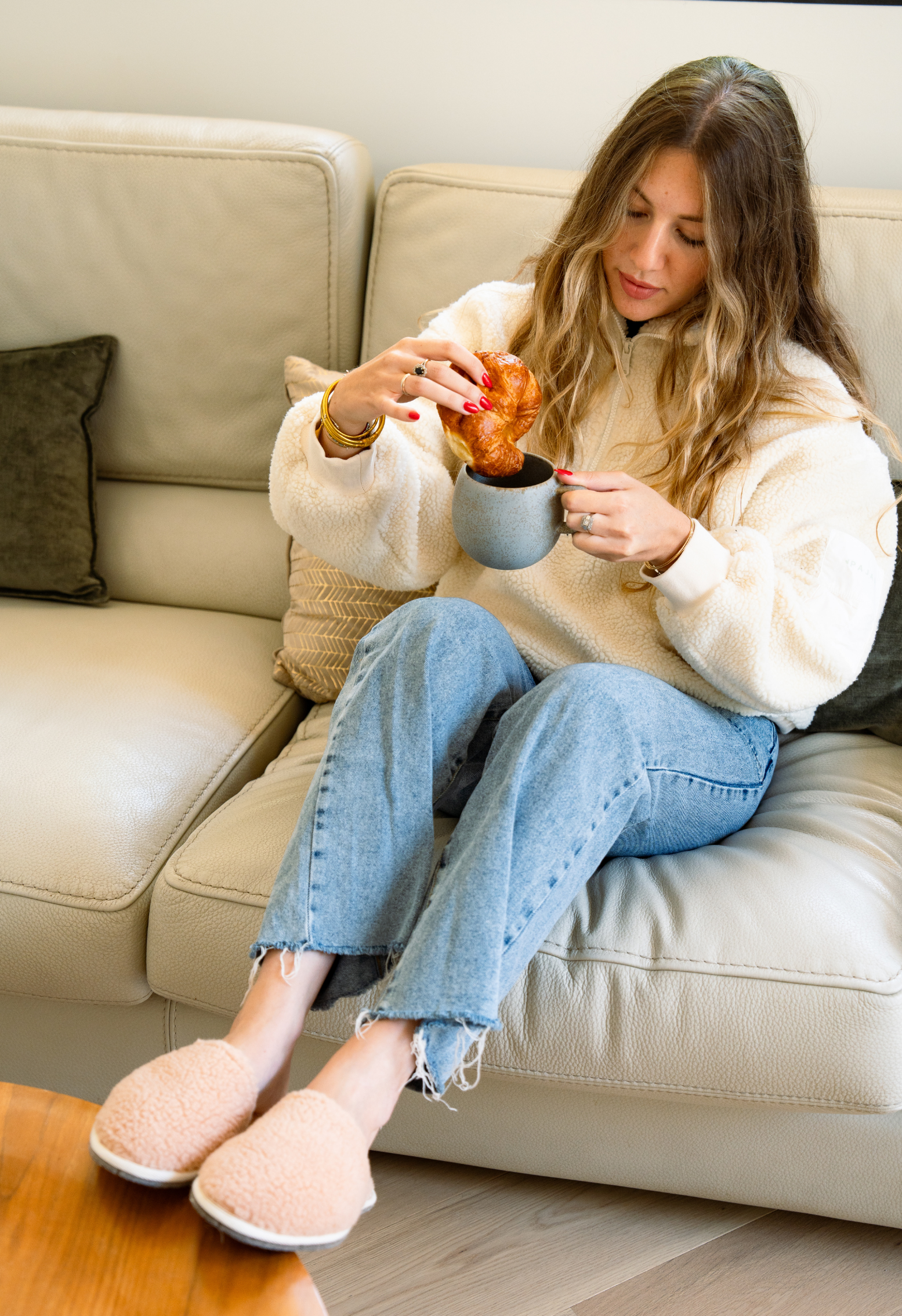 Femme déjeunant sur un canapé, trempant un croissant dans une tasse. Elle porte un pull en laine, un jean et des chaussons.