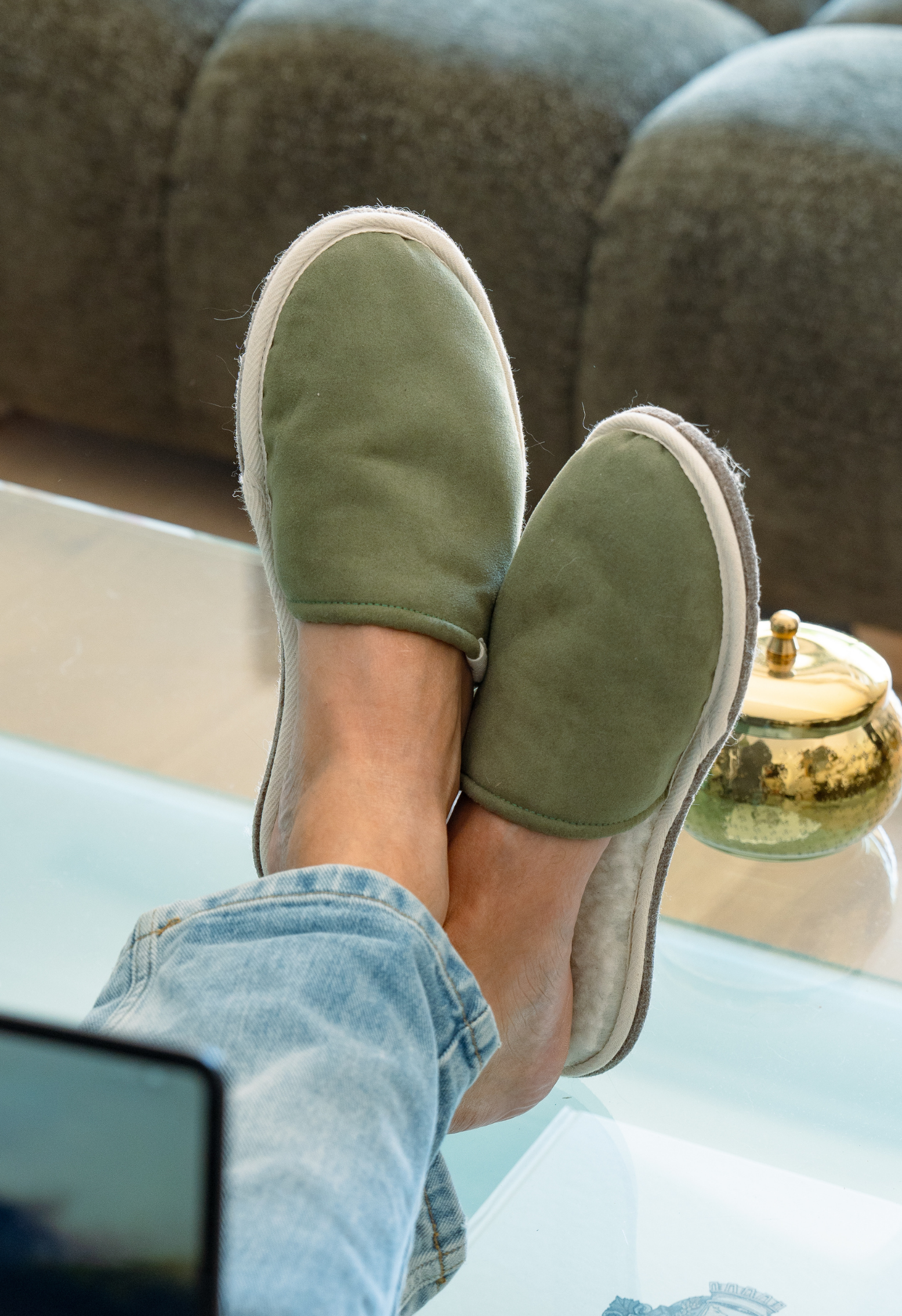 Pieds portant des pantoufles vertes sur une table basse en verre, une personne porte un jean bleu.