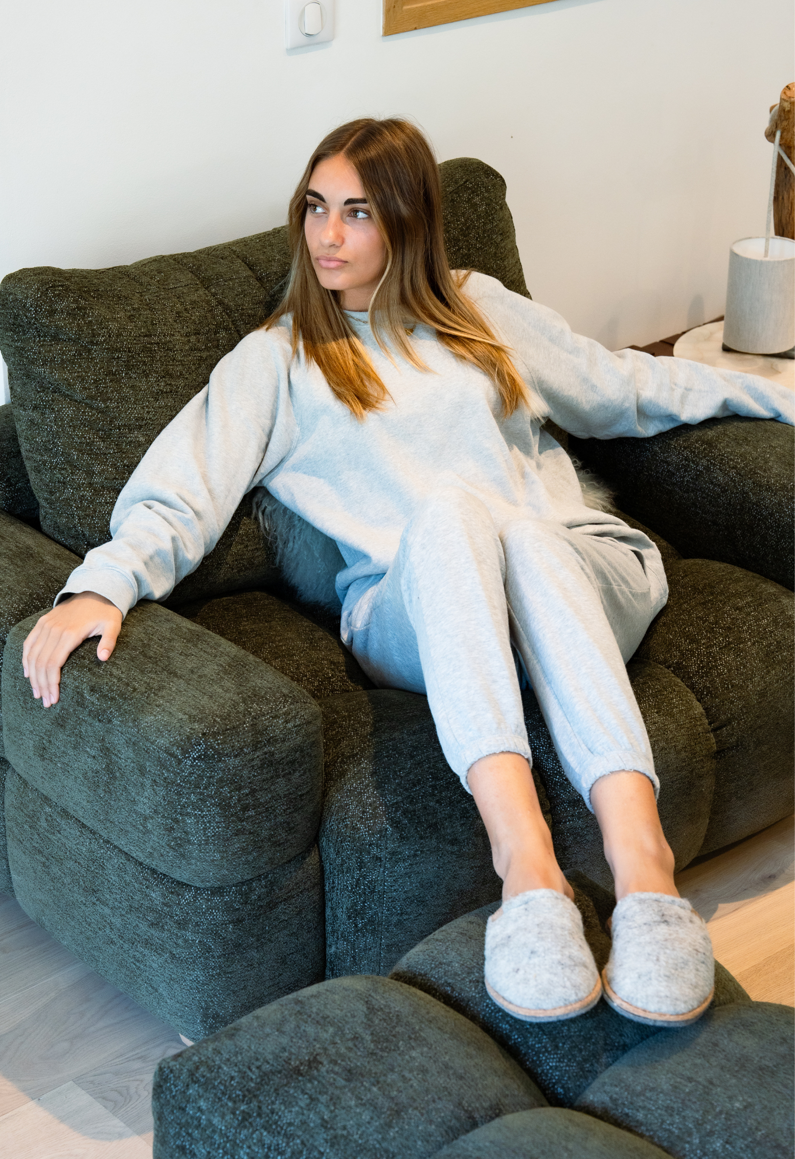 Jeune femme assise dans un fauteuil en tissu vert, vêtue d'un survêtement gris et de pantoufles, regardant sur le côté.