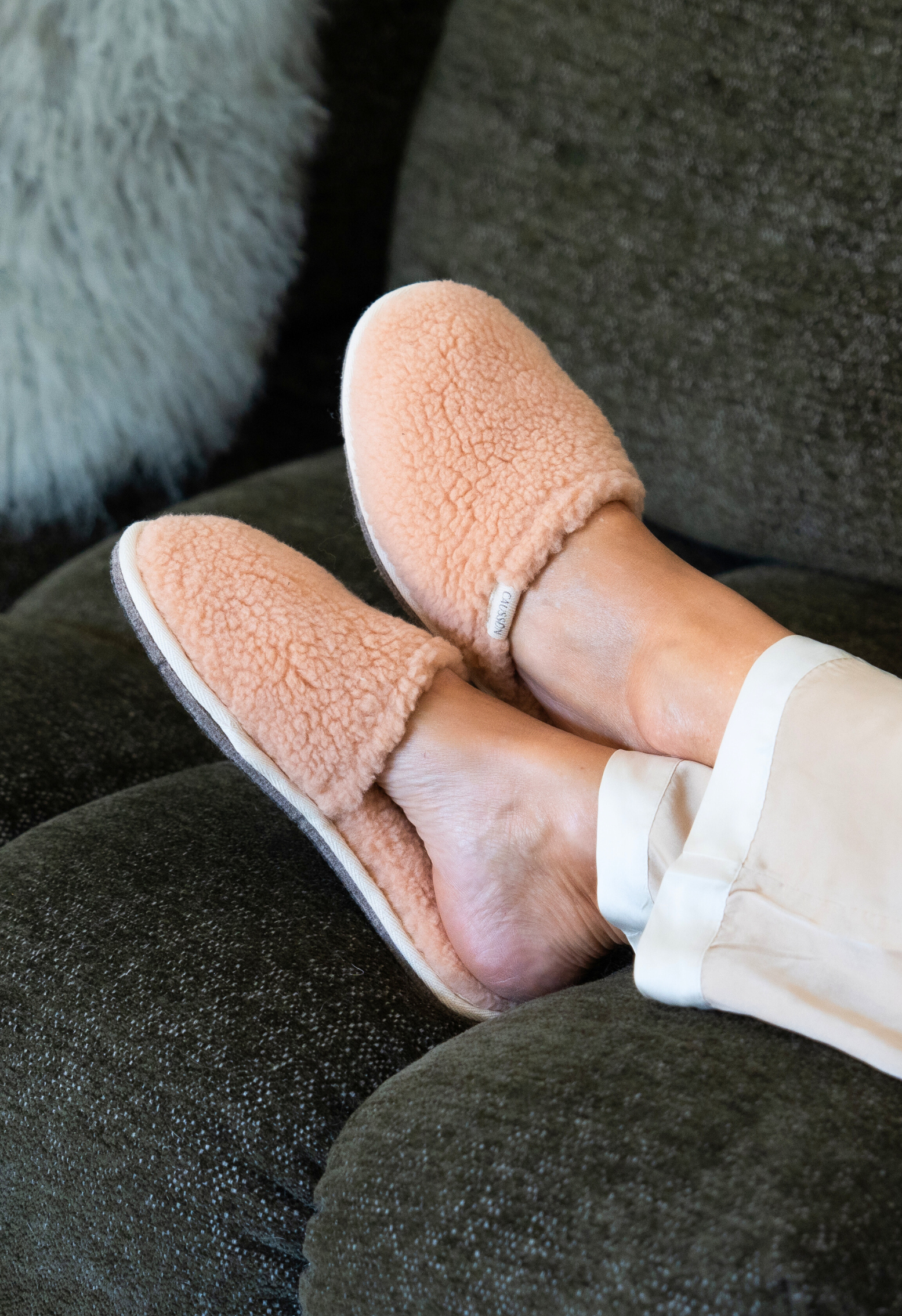 Gros plan des pieds portant des pantoufles en peau de mouton douillettes, posés sur un canapé vert foncé à coussins.