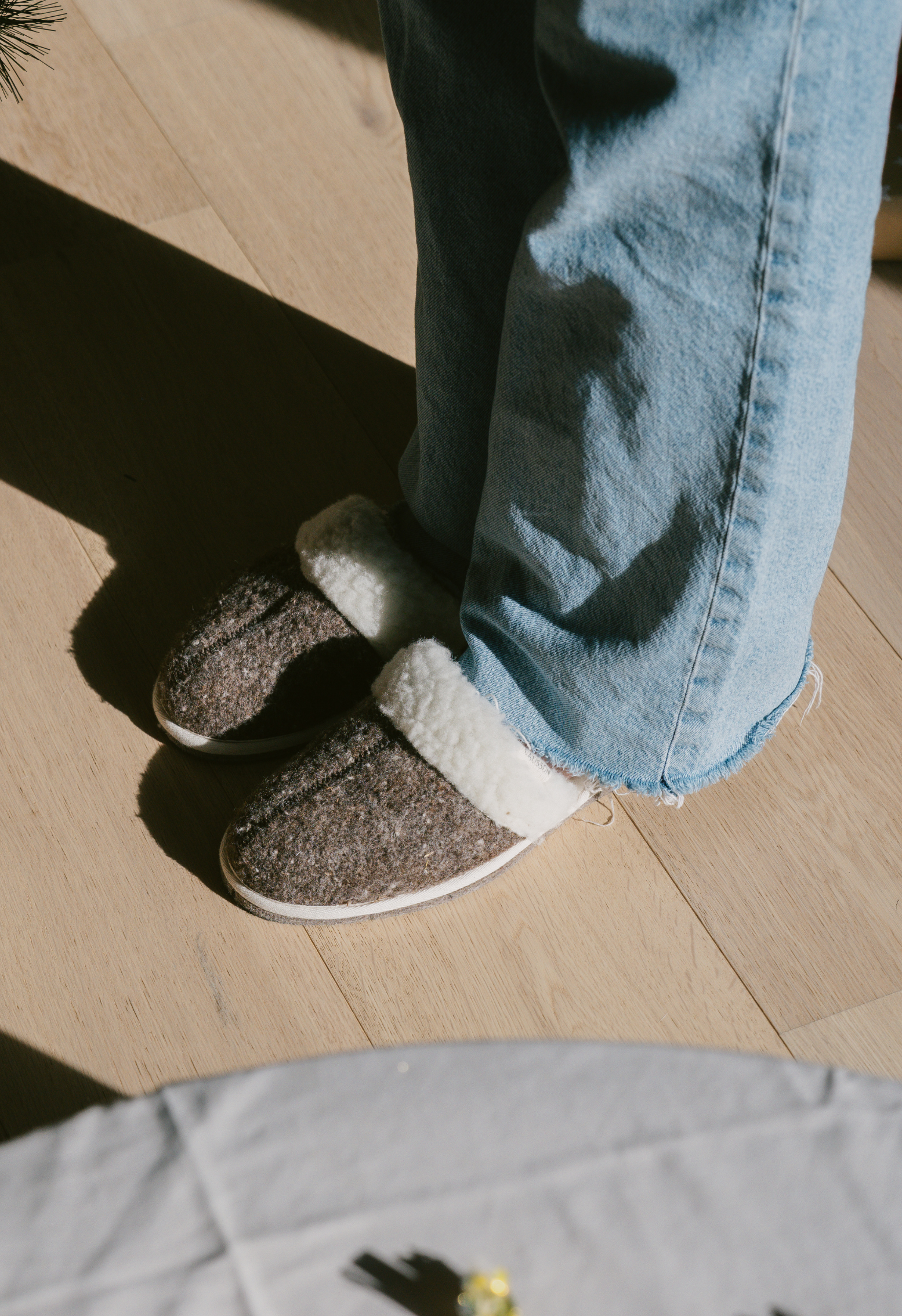 Une personne porte des pantoufles chaudes et douillettes. Elle porte un jean clair sur un plancher de bois franc baigné de soleil.