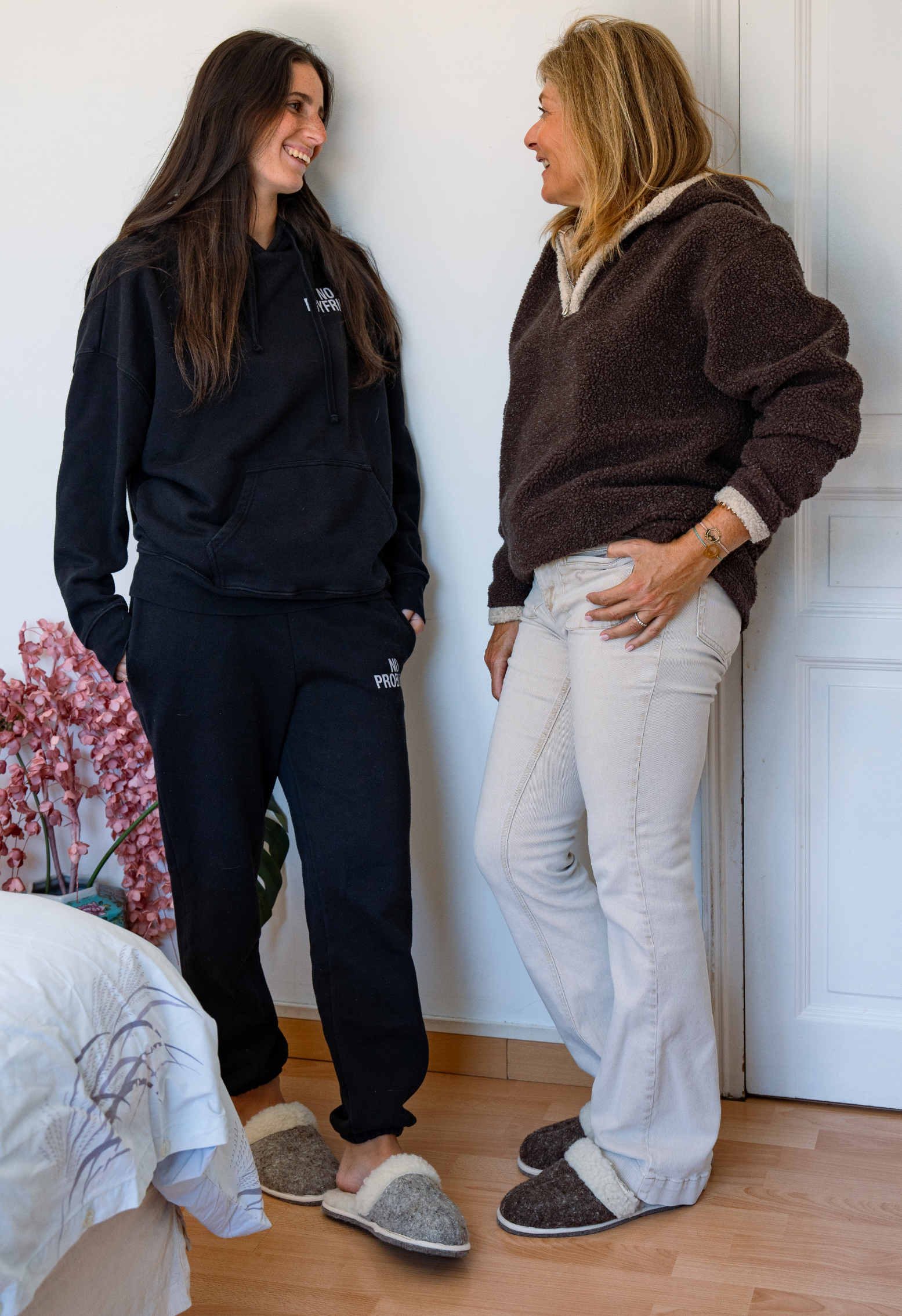 Deux femmes souriantes se regardent. L'une porte un survêtement noir, l'autre un sweat-shirt brun et un jean. Elles portent des chaussons.
