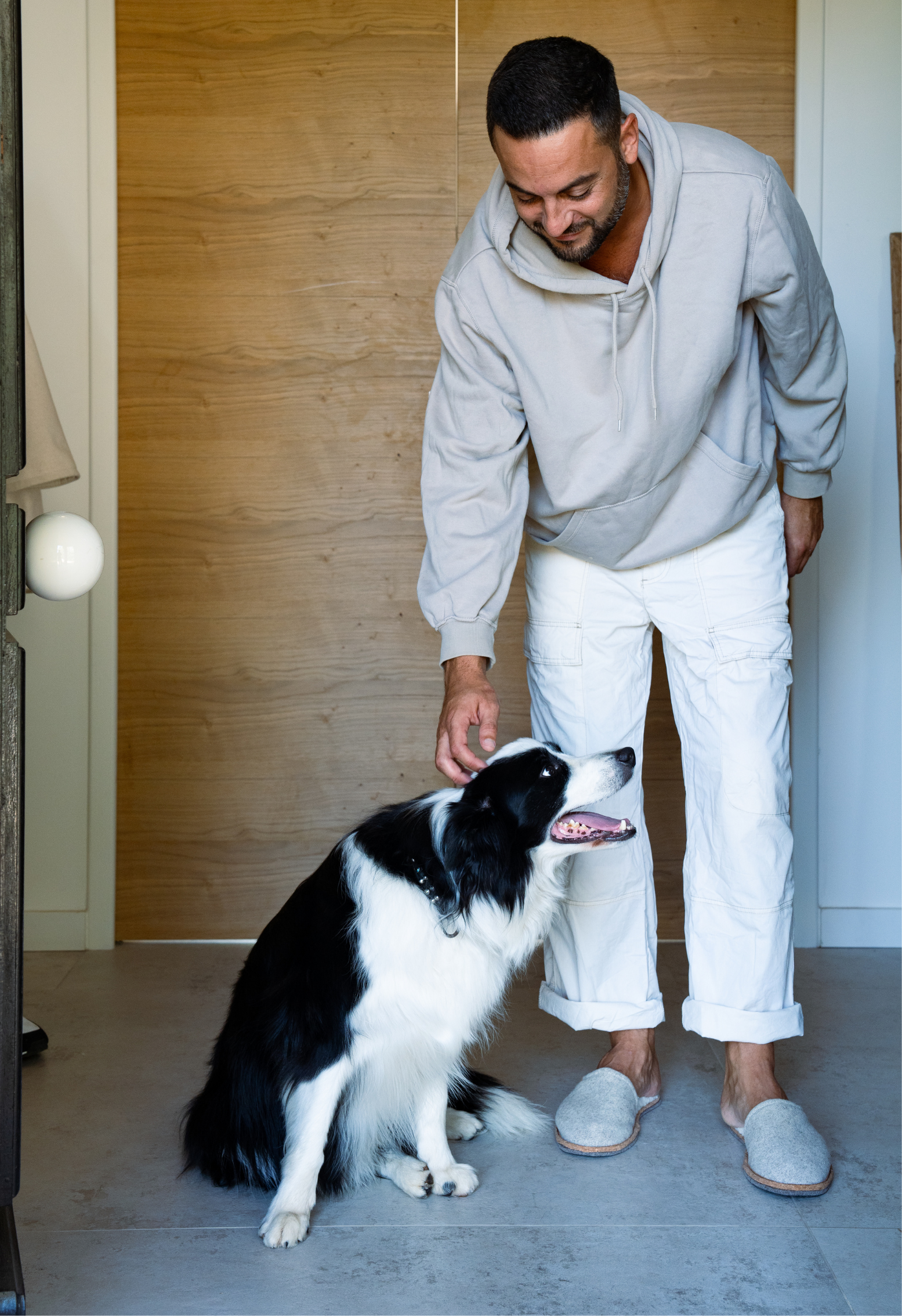 Un homme caresse un chien noir et blanc, dans une entrée moderne. L'homme porte un sweat à capuche et des chaussures pantoufles.