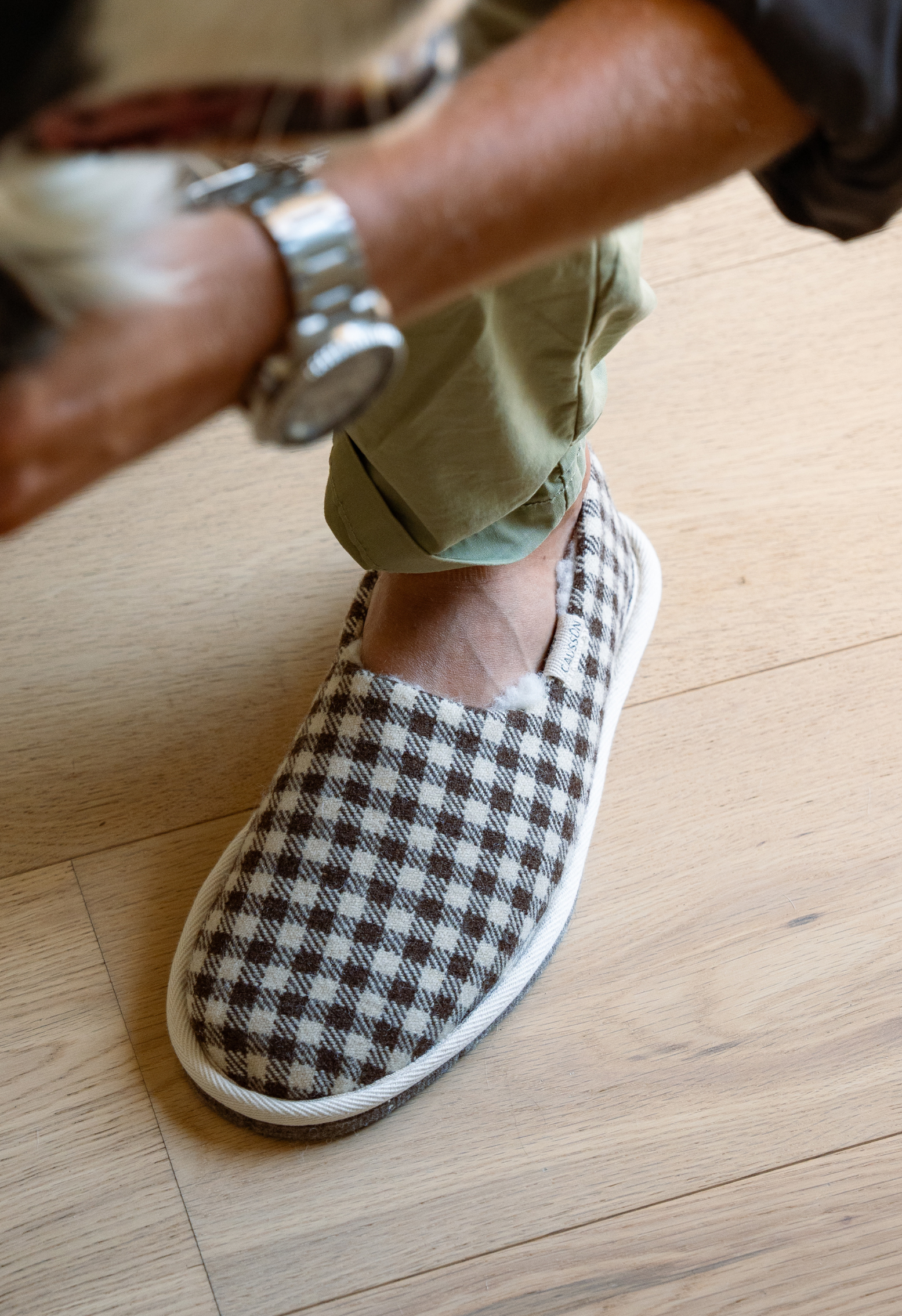 Gros plan sur un pied vêtu d'une pantoufle à carreaux marron et crème. Une personne porte une montre en argent et un pantalon vert.