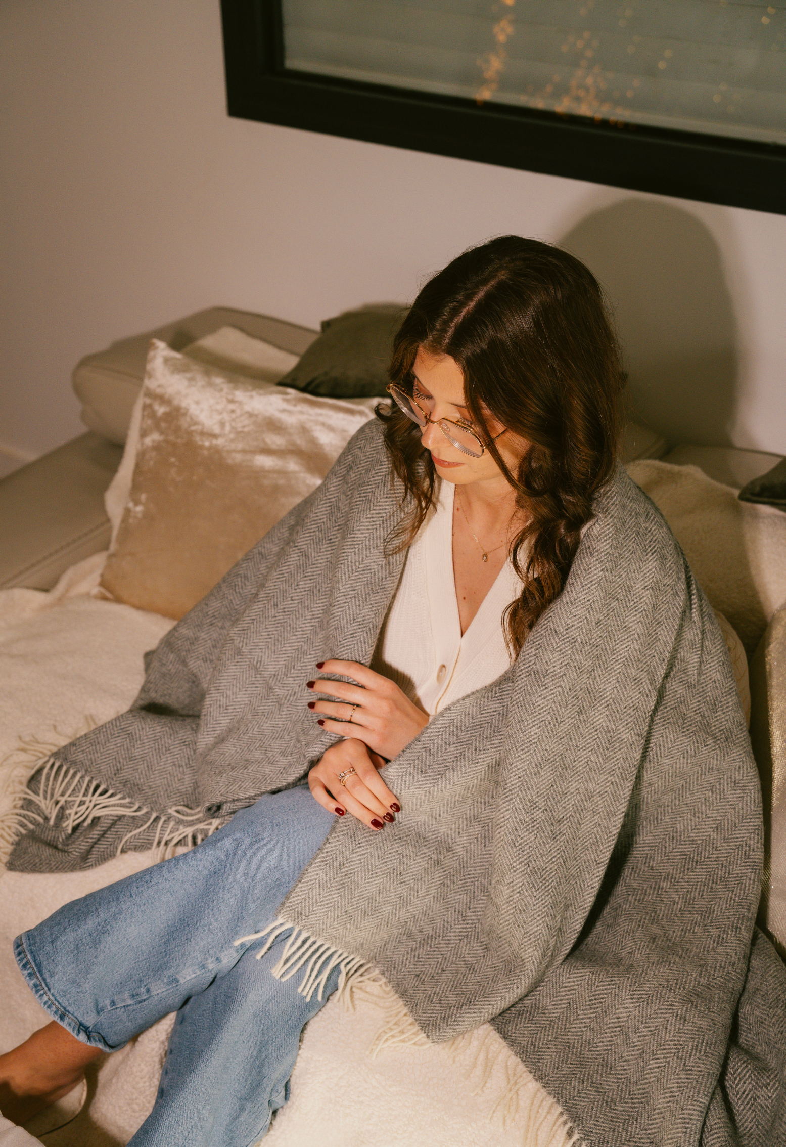 Femme portant des lunettes assise sur un canapé enveloppée dans une couverture grise à chevrons, jean bleu, ambiance cosy et détendue.