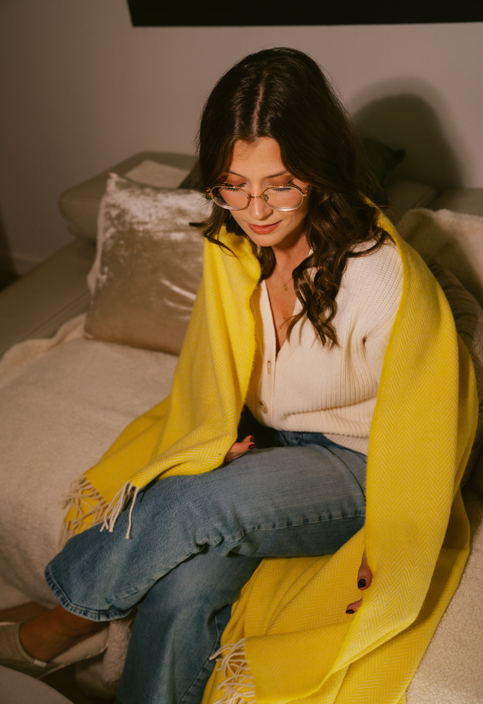 Jeune femme assise sur un canapé enveloppée dans une couverture jaune. Elle porte des lunettes, un jean et un pull blanc.