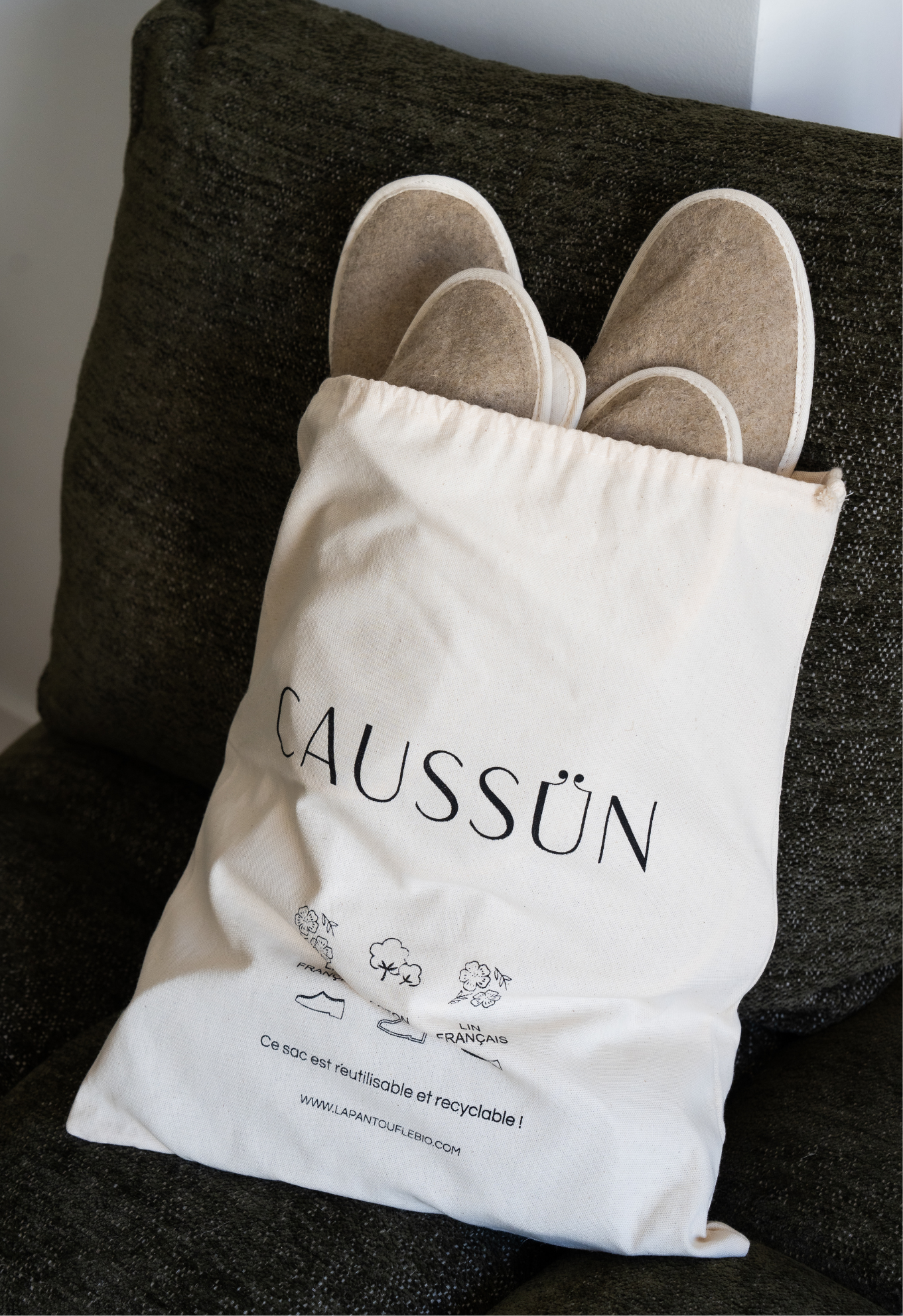 Un sac en coton avec le logo CAUSSUN contient des chaussons en laine beige, posé sur un coussin vert foncé. Le sac est réutilisable et recyclable.