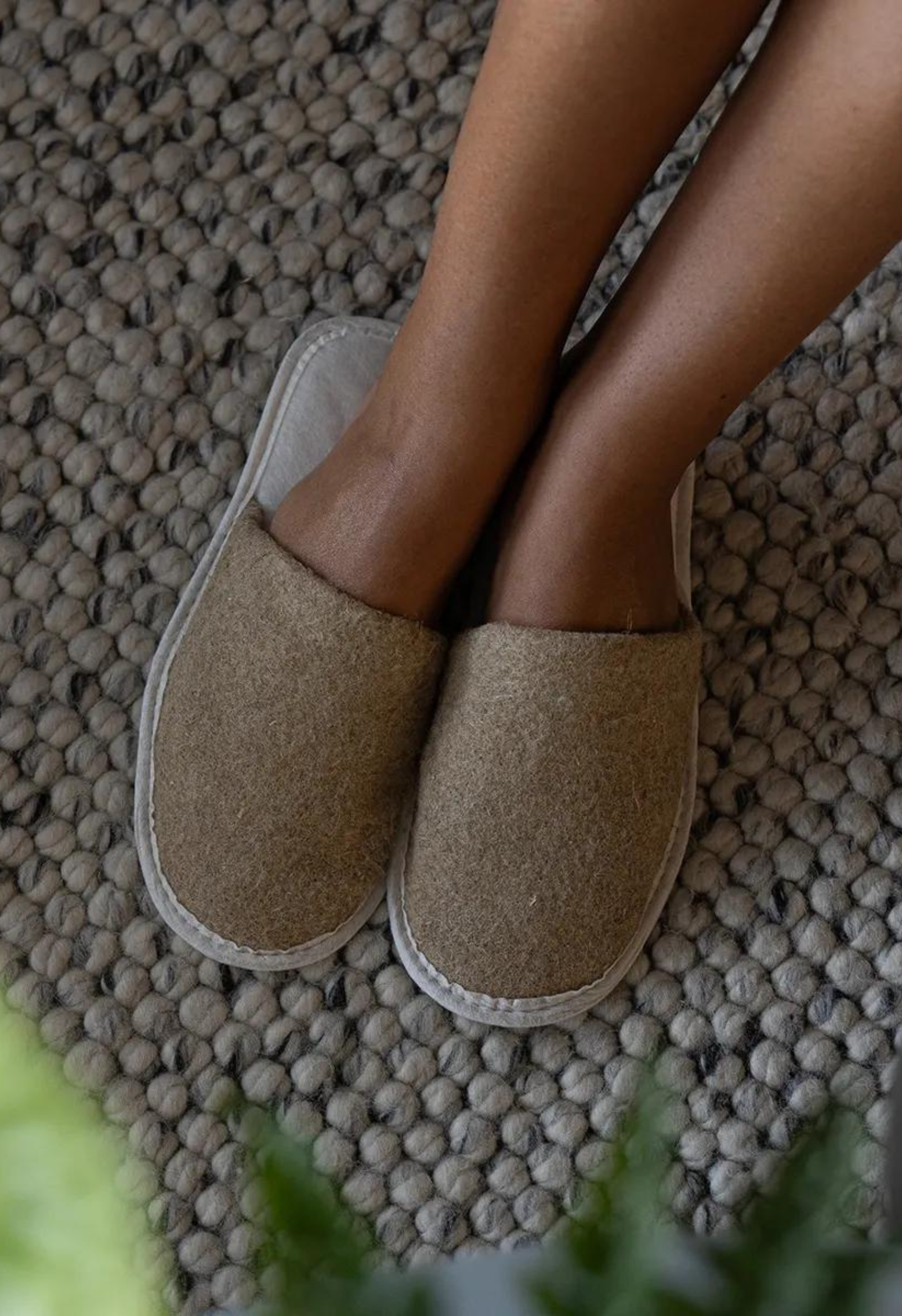 Gros plan des pieds dans des pantoufles en laine, sur un tapis texturé. Le tapis est fait de boules de laine grises.