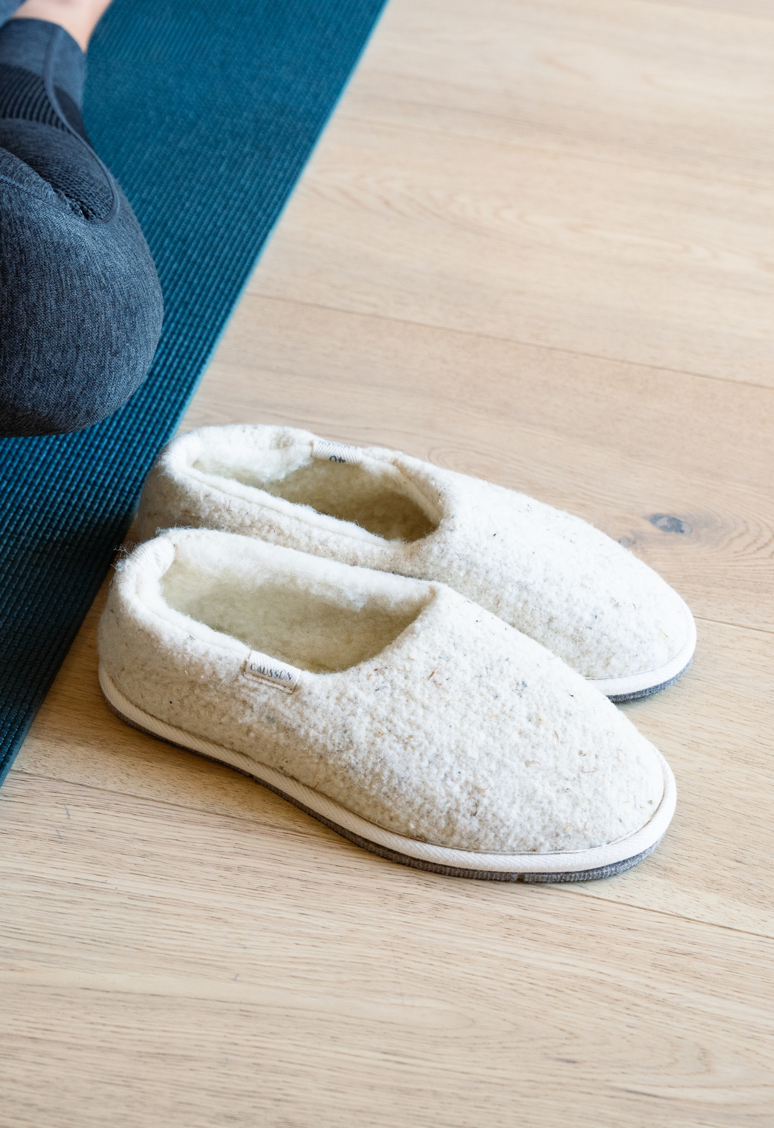 Paire de pantoufles en laine blanche sur un parquet en bois, à côté d'un tapis de yoga bleu et d'une personne assise.