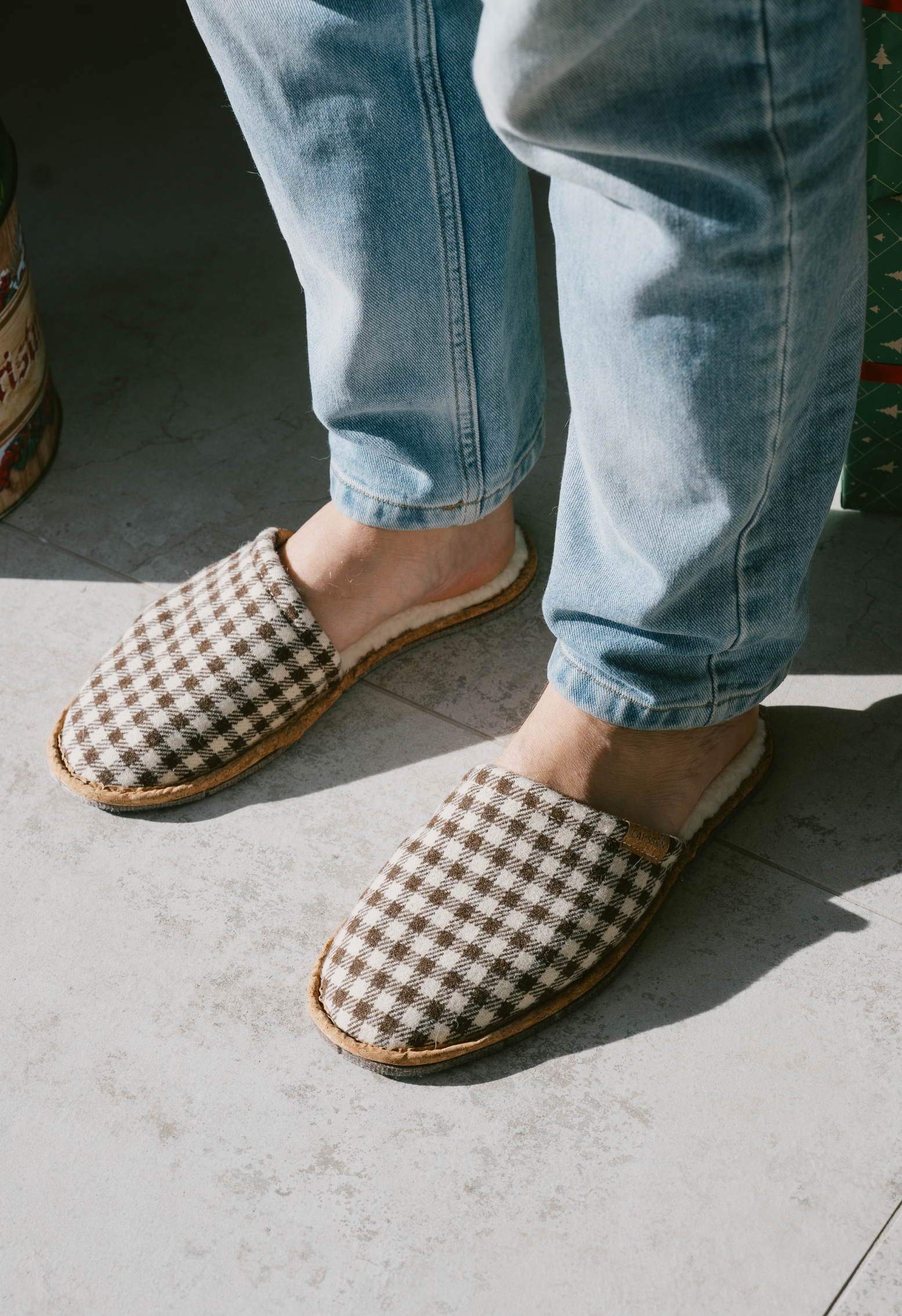 Gros plan des pieds avec des pantoufles à carreaux marron et des jeans bleus sur un sol en béton gris clair.