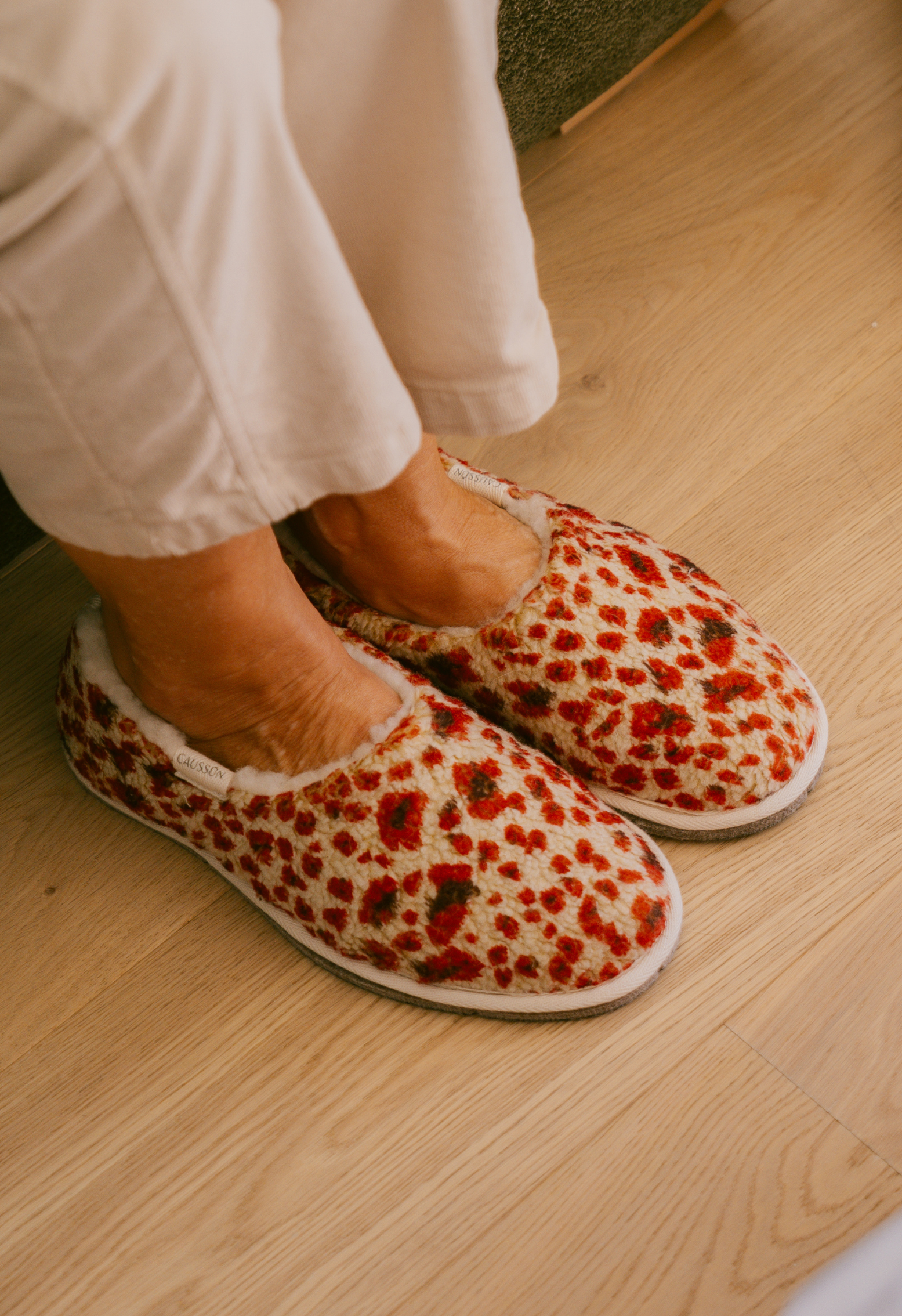 Gros plan de pantoufles imprimées léopard avec des pieds en pantalon blanc sur un parquet en bois.