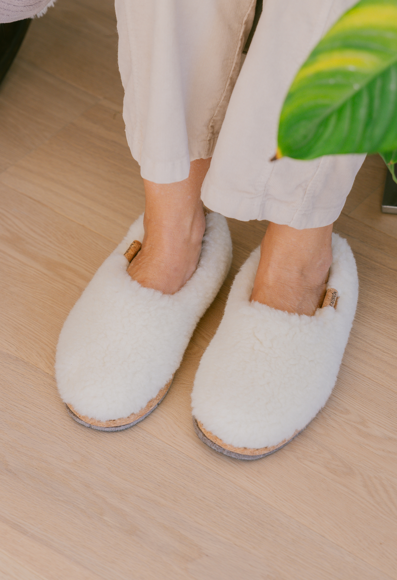 Gros plan de pieds portant des pantoufles en laine blanche et un pantalon en lin beige sur un plancher de bois. Une plante verte sur le côté.