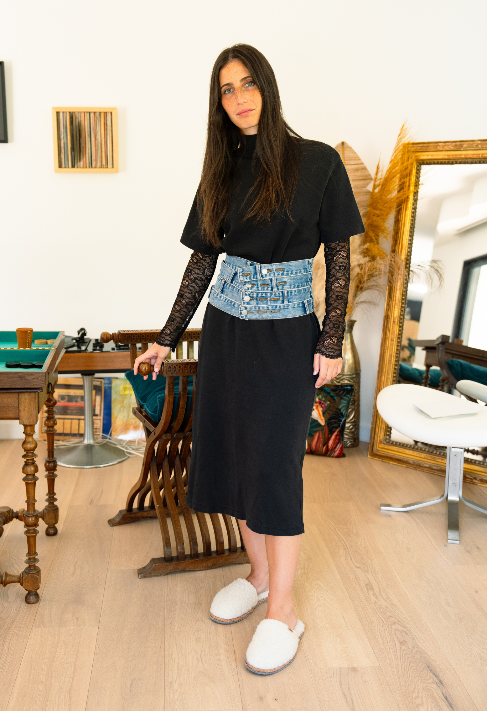 Une femme vêtue d'une robe noire avec une ceinture en jean et des chaussons, posant dans un intérieur moderne avec miroir.