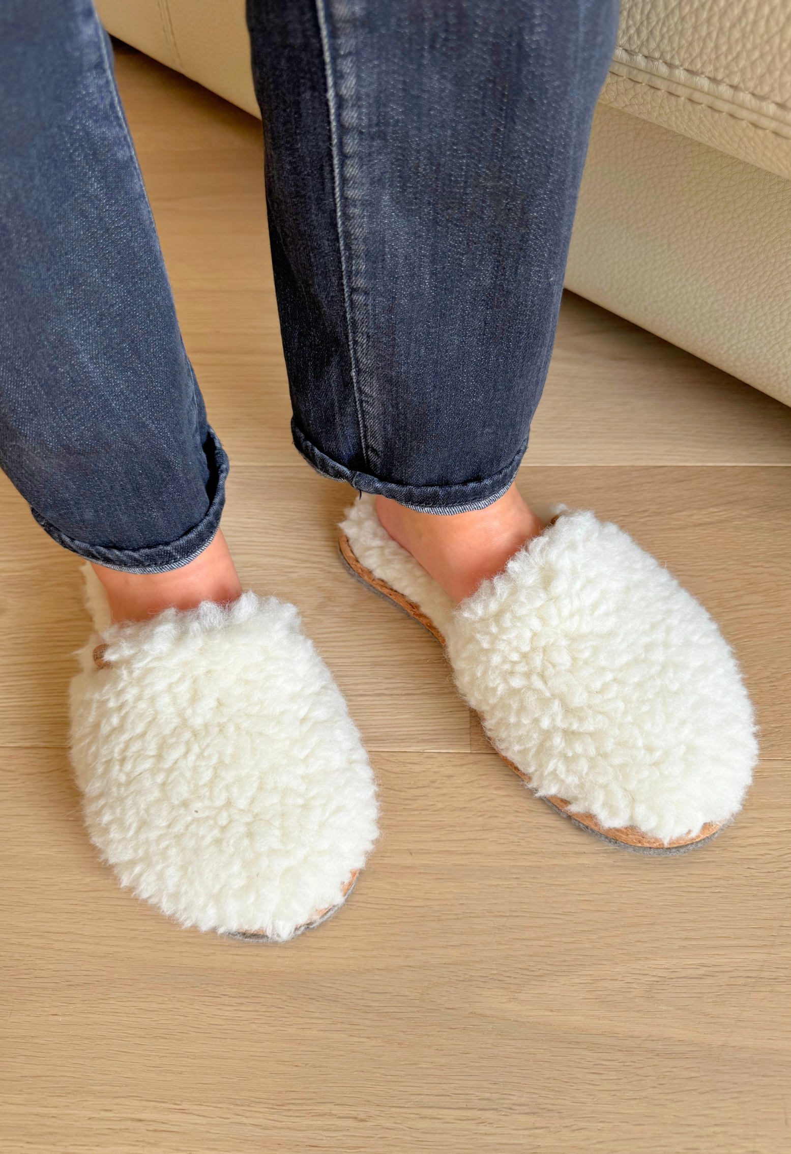 Pieds dans des chaussons en laine de mouton, portant un jean bleu, sur un plancher en bois clair, près d'un canapé crème.